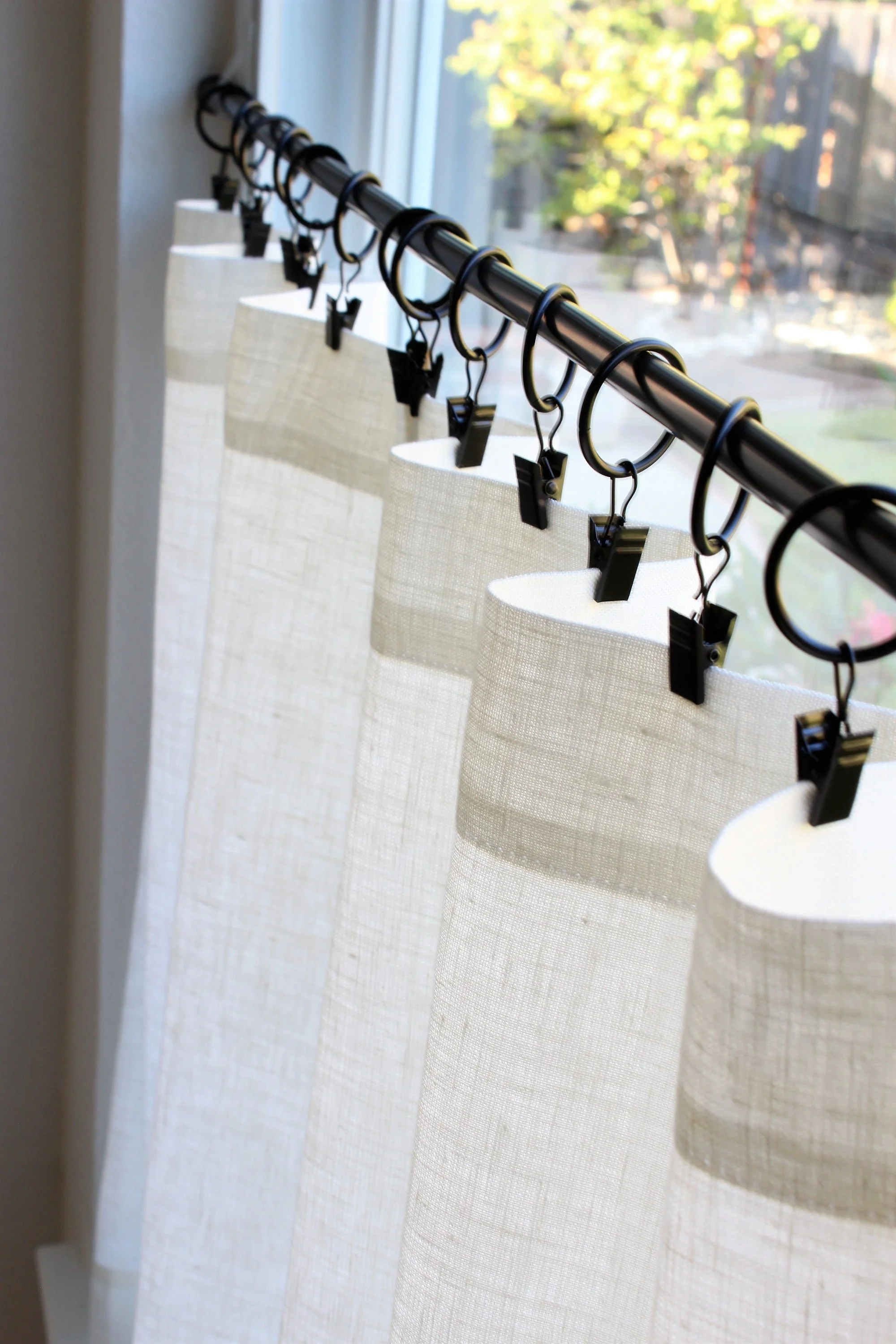 A row of white curtains hanging from a black curtain rod with clips, by a window with trees outside.