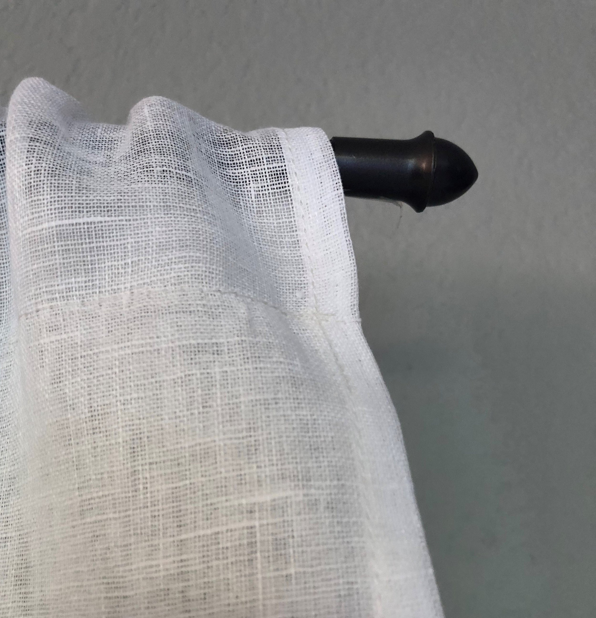 Close-up image of a white sheer curtain hanging from a black curtain rod against a gray background.