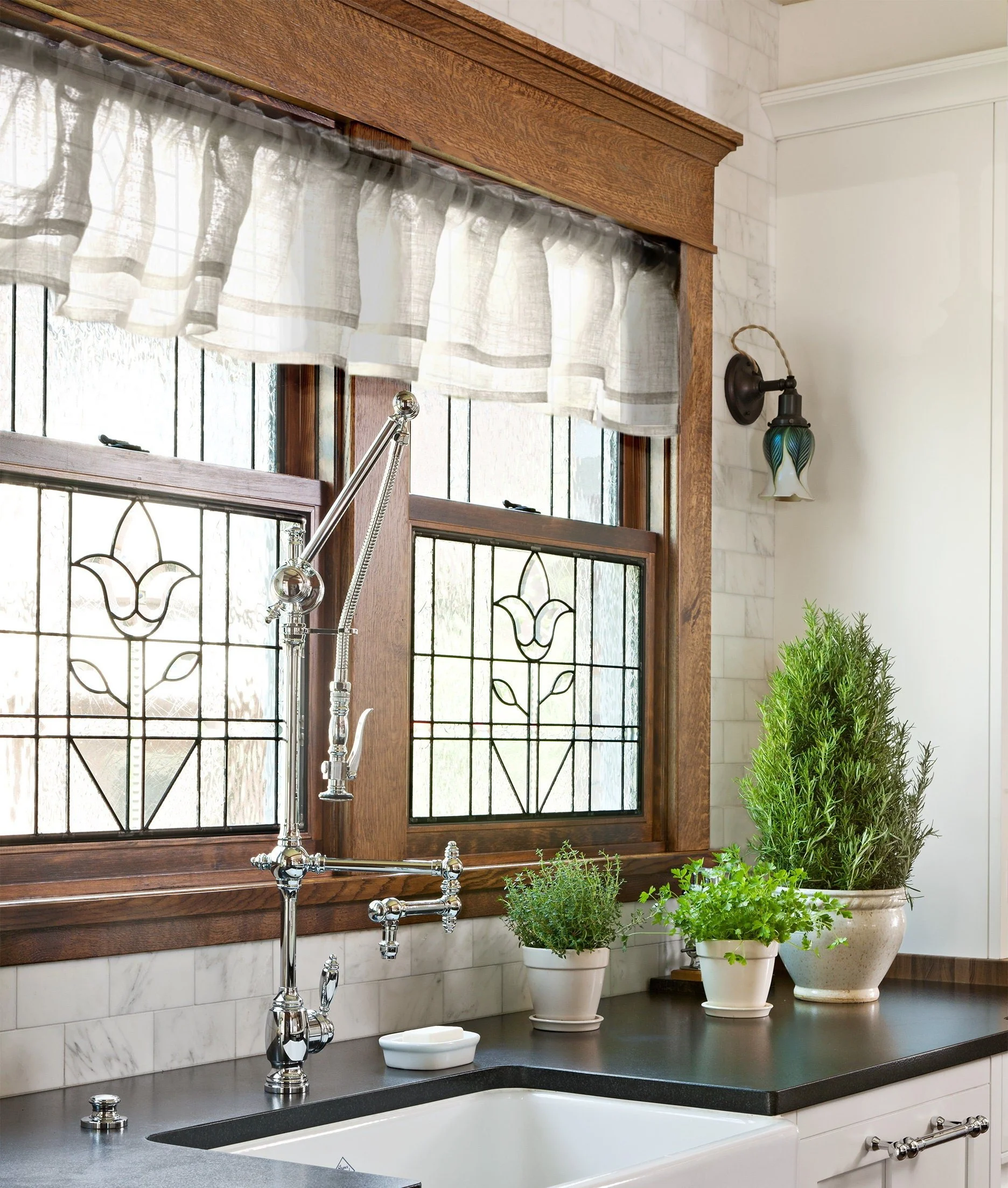 Kitchen window with stained glass design and curtains, wood trim, plants on black countertop, chrome kitchen faucet, and a wall sconce light.