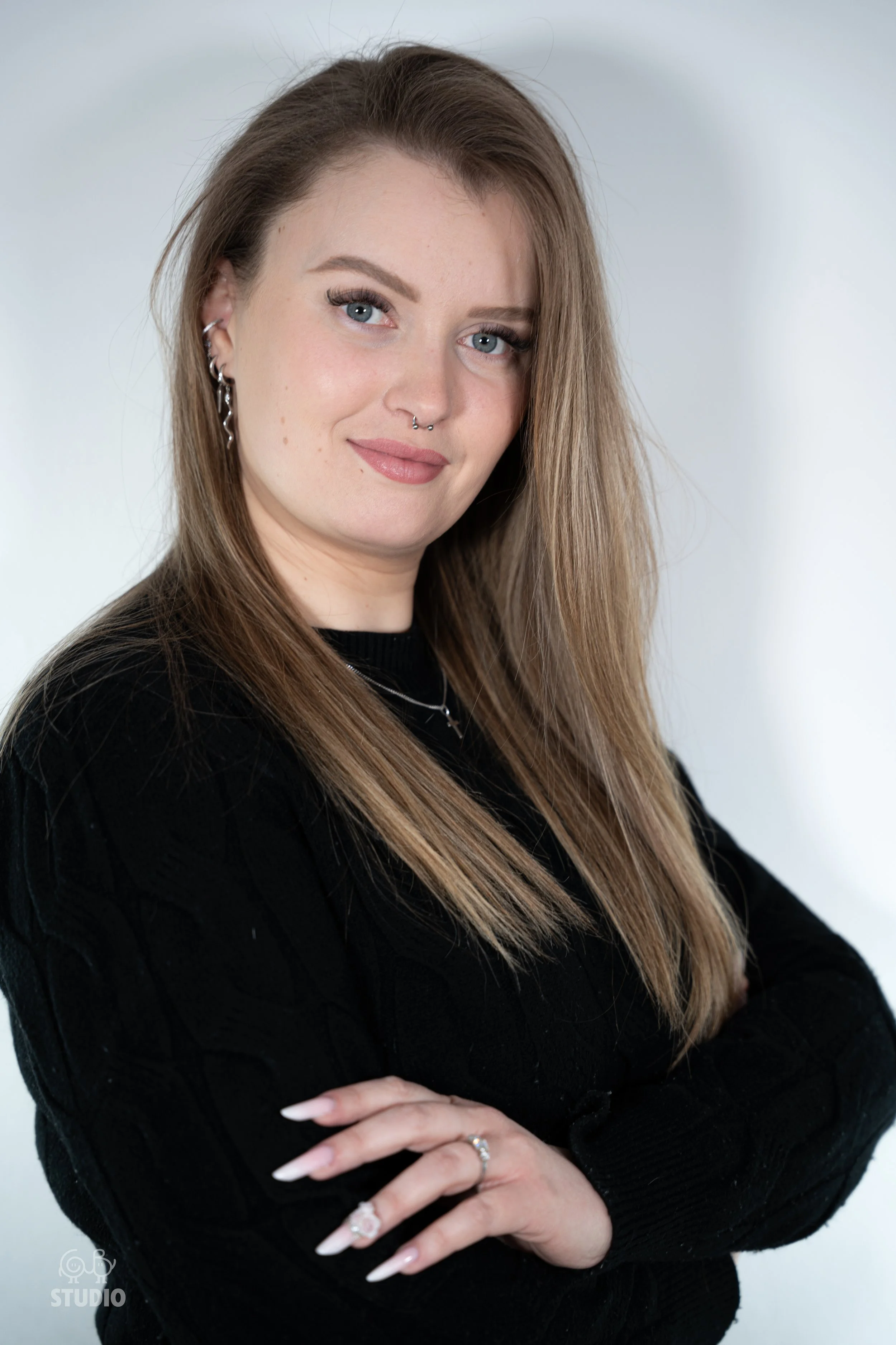 Perrine, la prothésiste ongulaire aux cheveux longs et blonds, portant un pull noir, avec des boucles d'oreilles, un piercing au nez et un sourire léger, croise les bras debout devant un fond blanc.