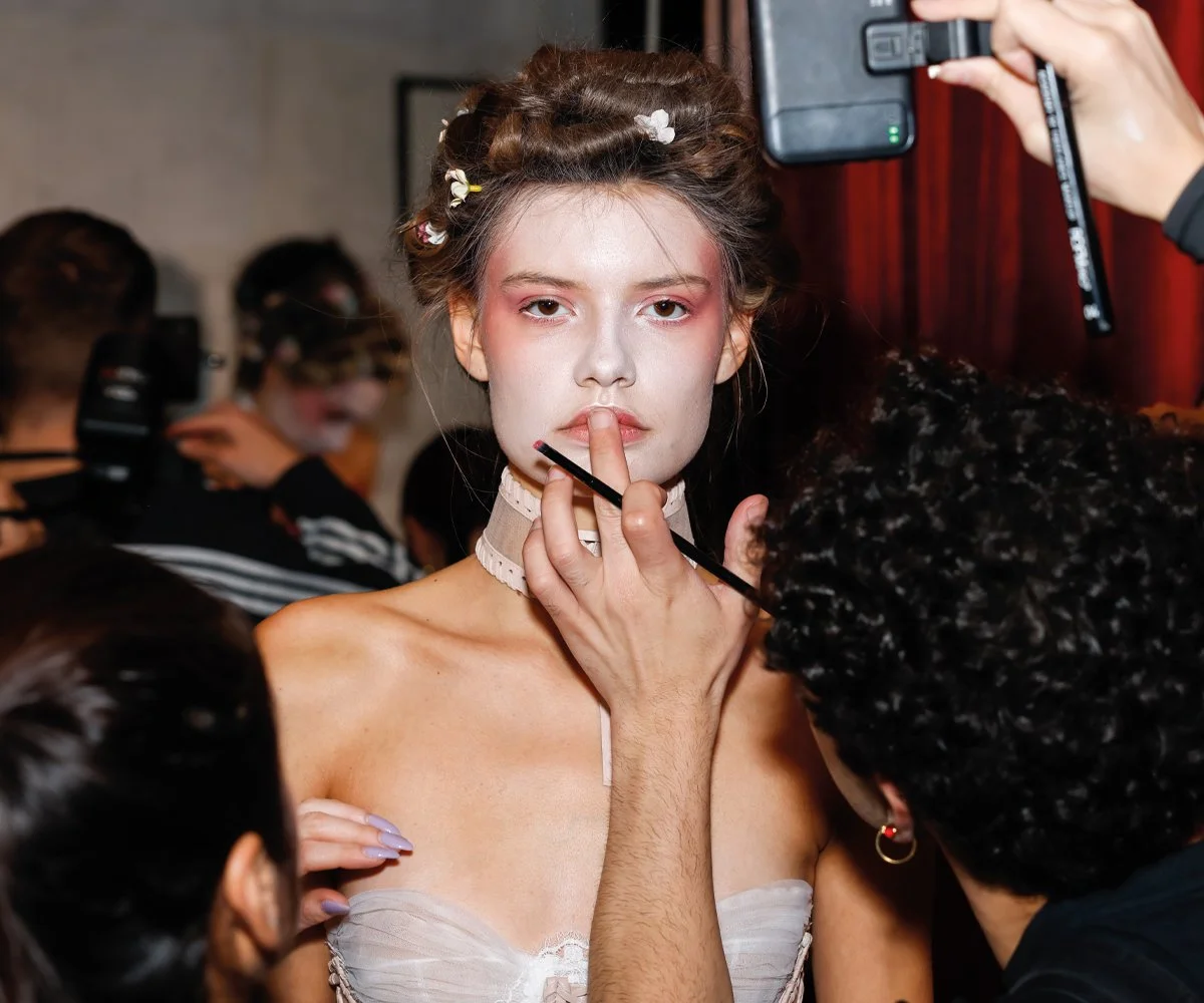 Editorial behind-the-scenes fashion photograph showing hair and makeup preparation backstage