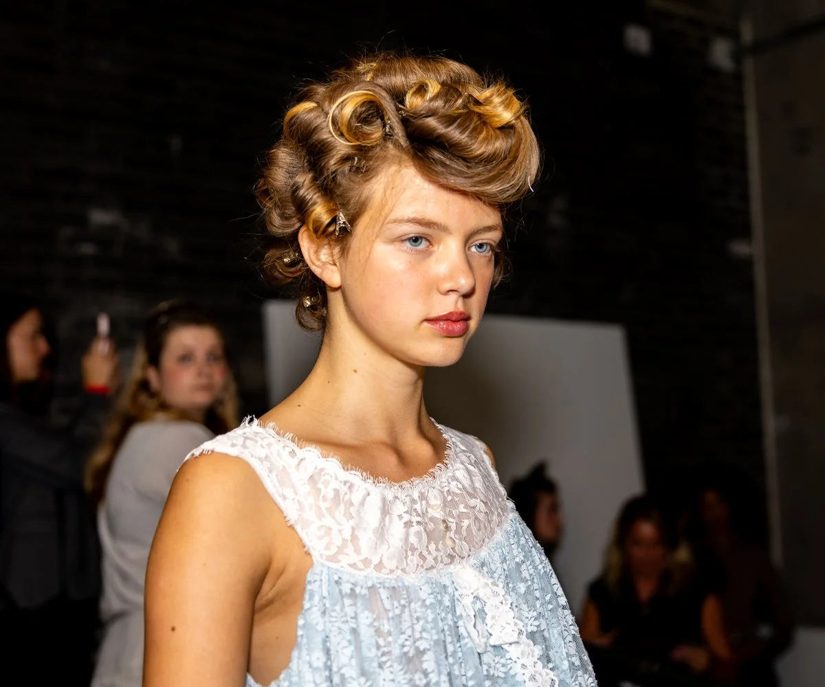 Model backstage at London Fashion Week wearing a pale blue lace dress with vintage-inspired hair.