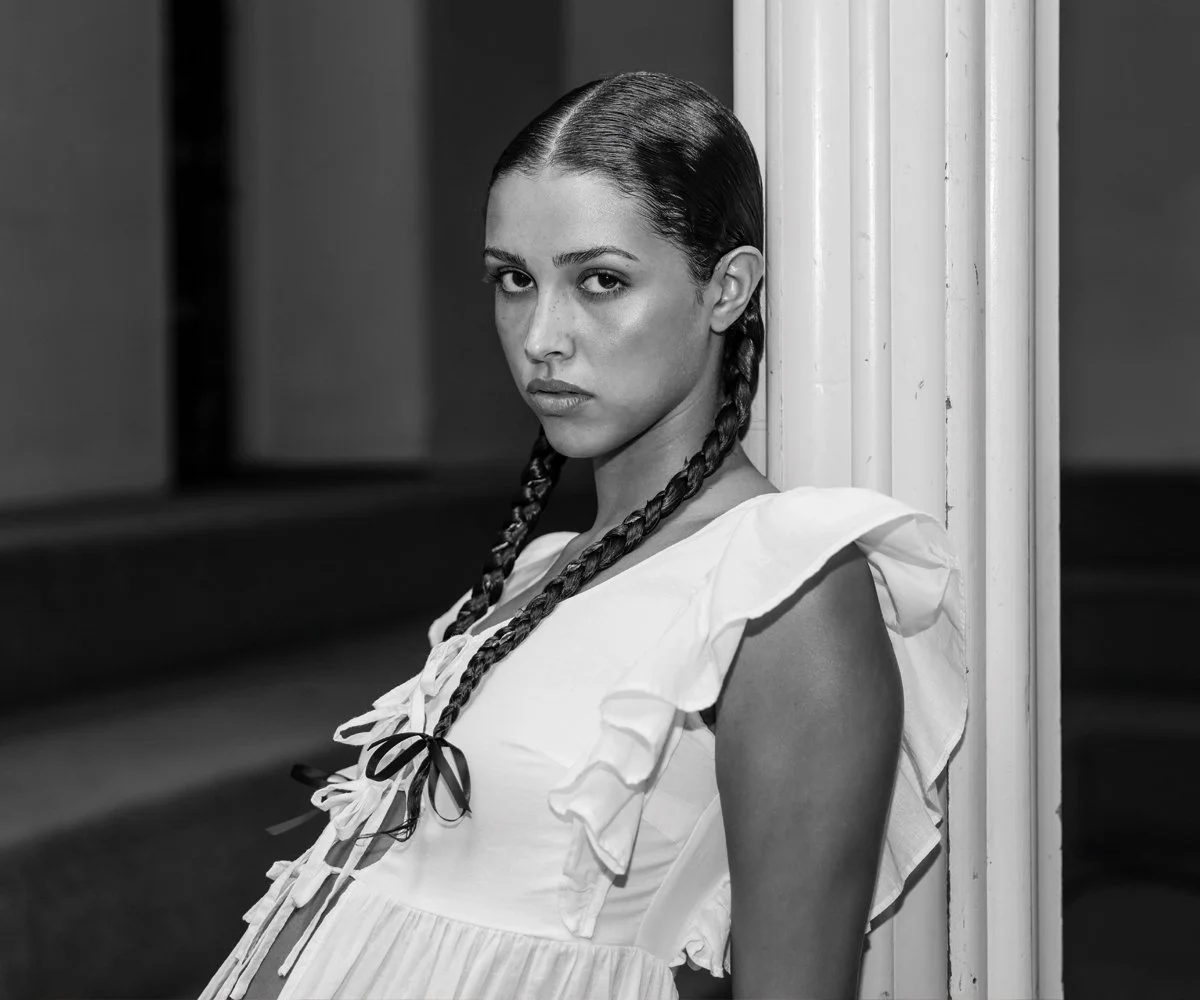 A black-and-white photo of a young woman with slicked-back hair styled in two braids, wearing a ruffled sleeveless dress, standing against a decorative column and looking directly at the camera.