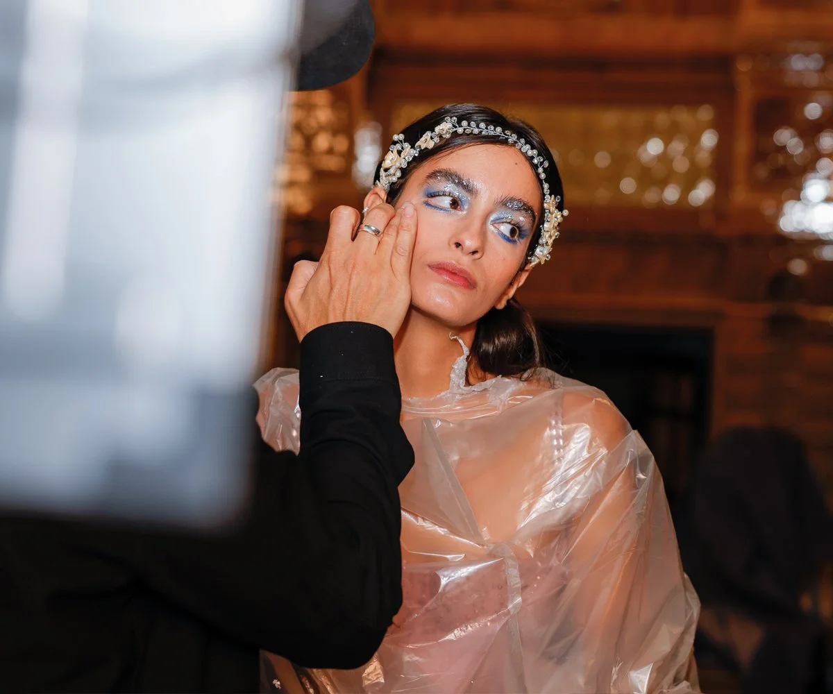 A woman with blue eye makeup and a jeweled headband is getting her makeup done by a person preparing her for an event or photo shoot.
