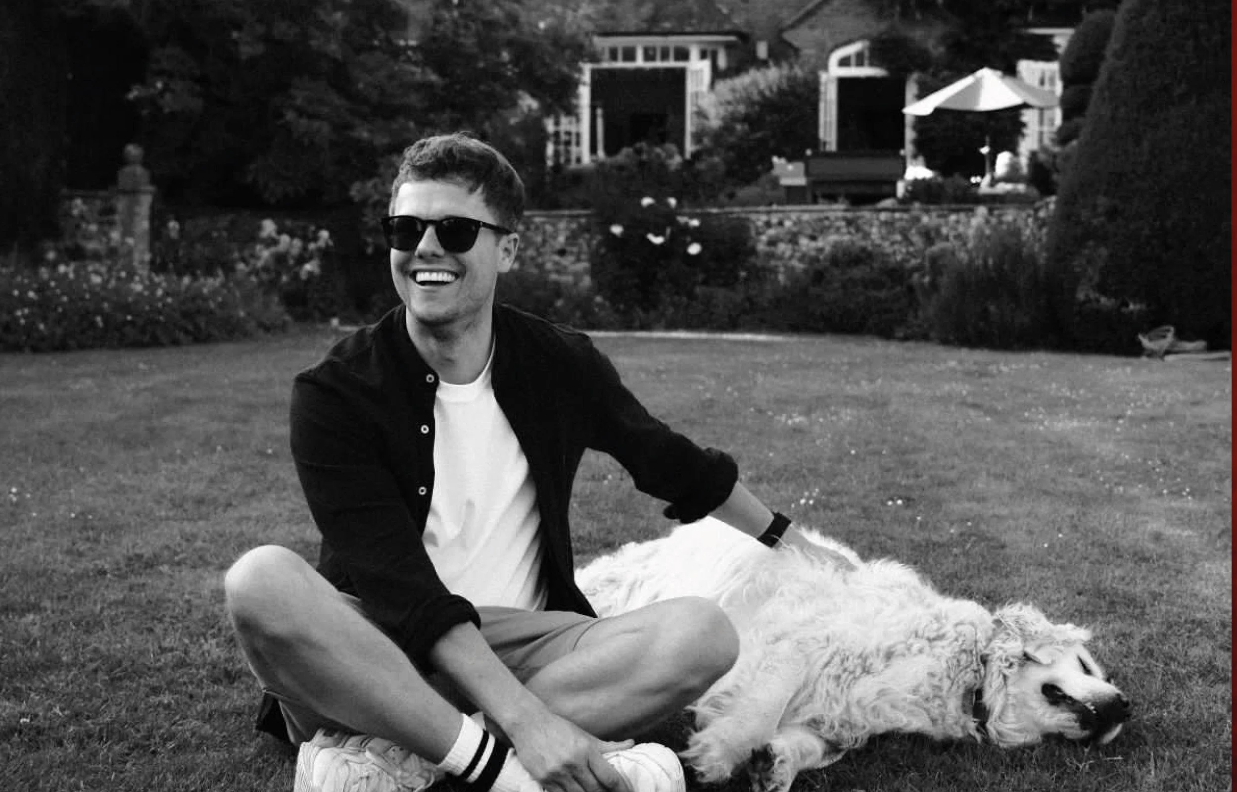A young man with short hair wearing sunglasses, a white t-shirt, and a black shirt, sitting on the grass and smiling, with a relaxed dog lying beside him, in a garden with trees, flowers, and large buildings in the background.