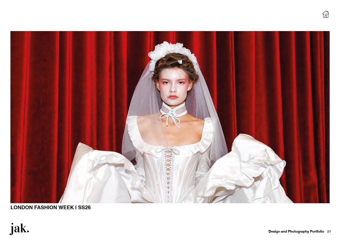 A female model on a runway dressed in a white bridal gown with ruffled details, lace-up corset, and veil, standing in front of red curtains.