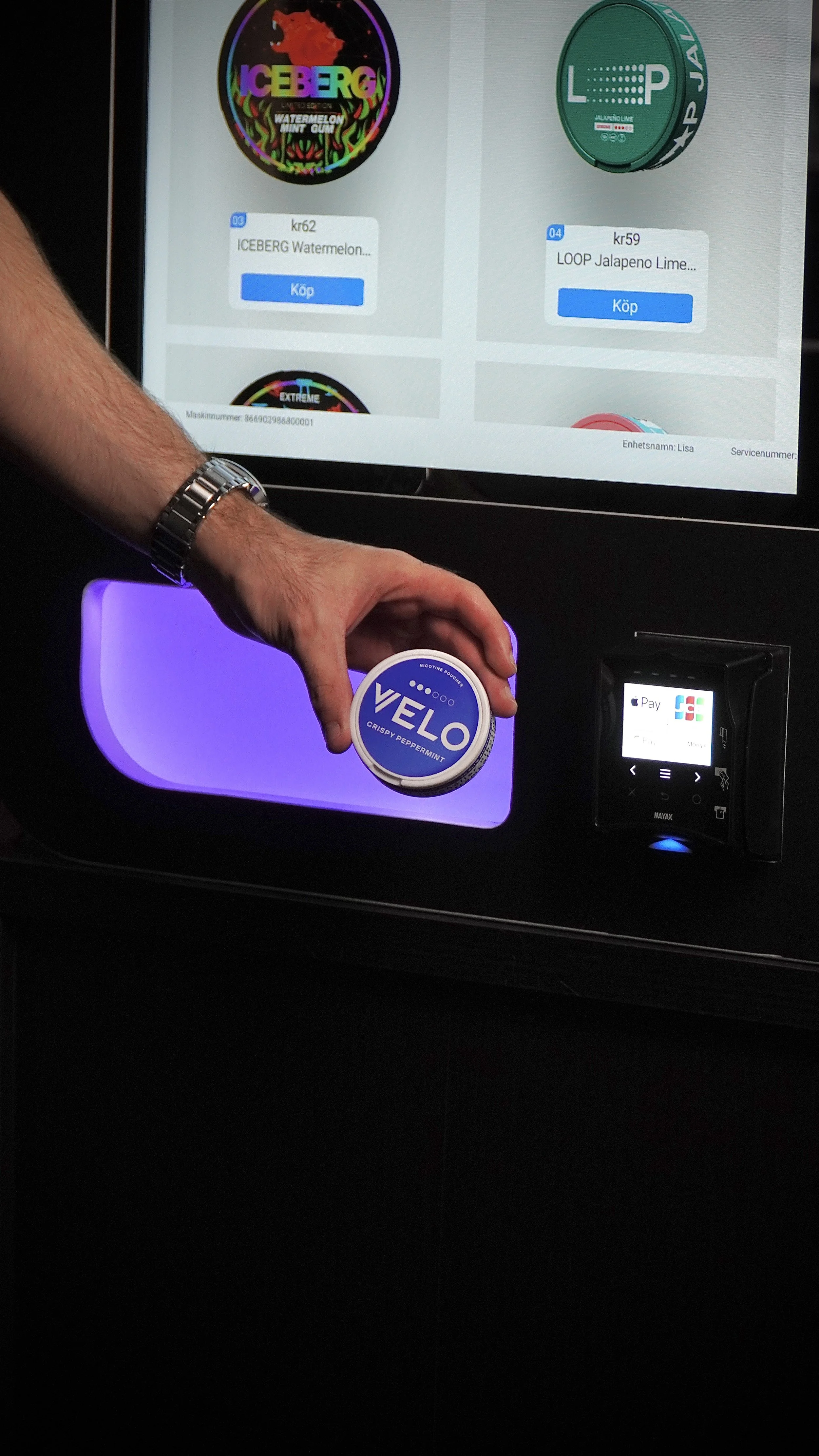 En person håller i ett snus med Velo-kapsel, framför en skärm med olika produktannonser på ett kafé eller automat.