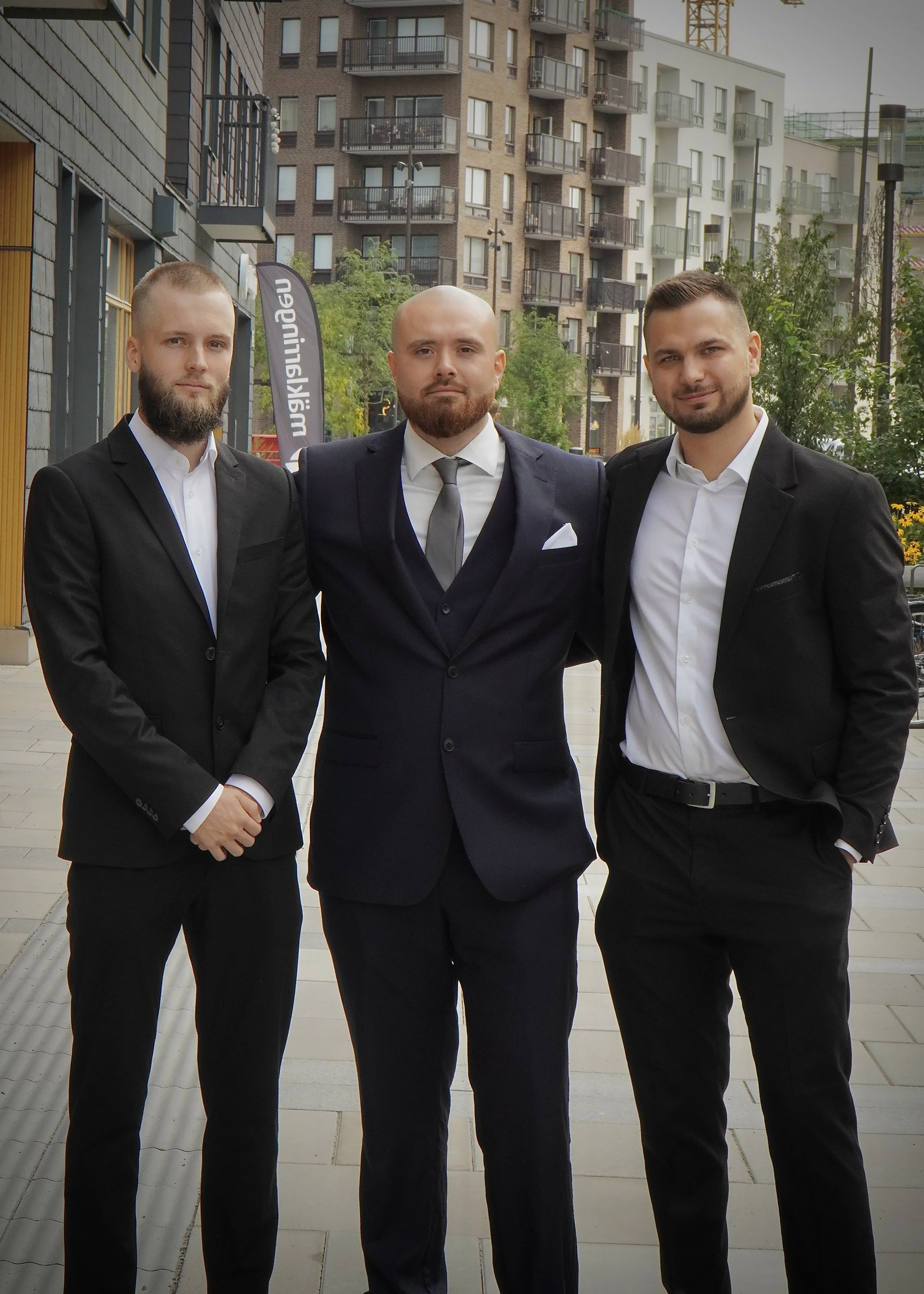 Tre män i kostymer står utomhus framför moderna bostadshus, pose för foto.