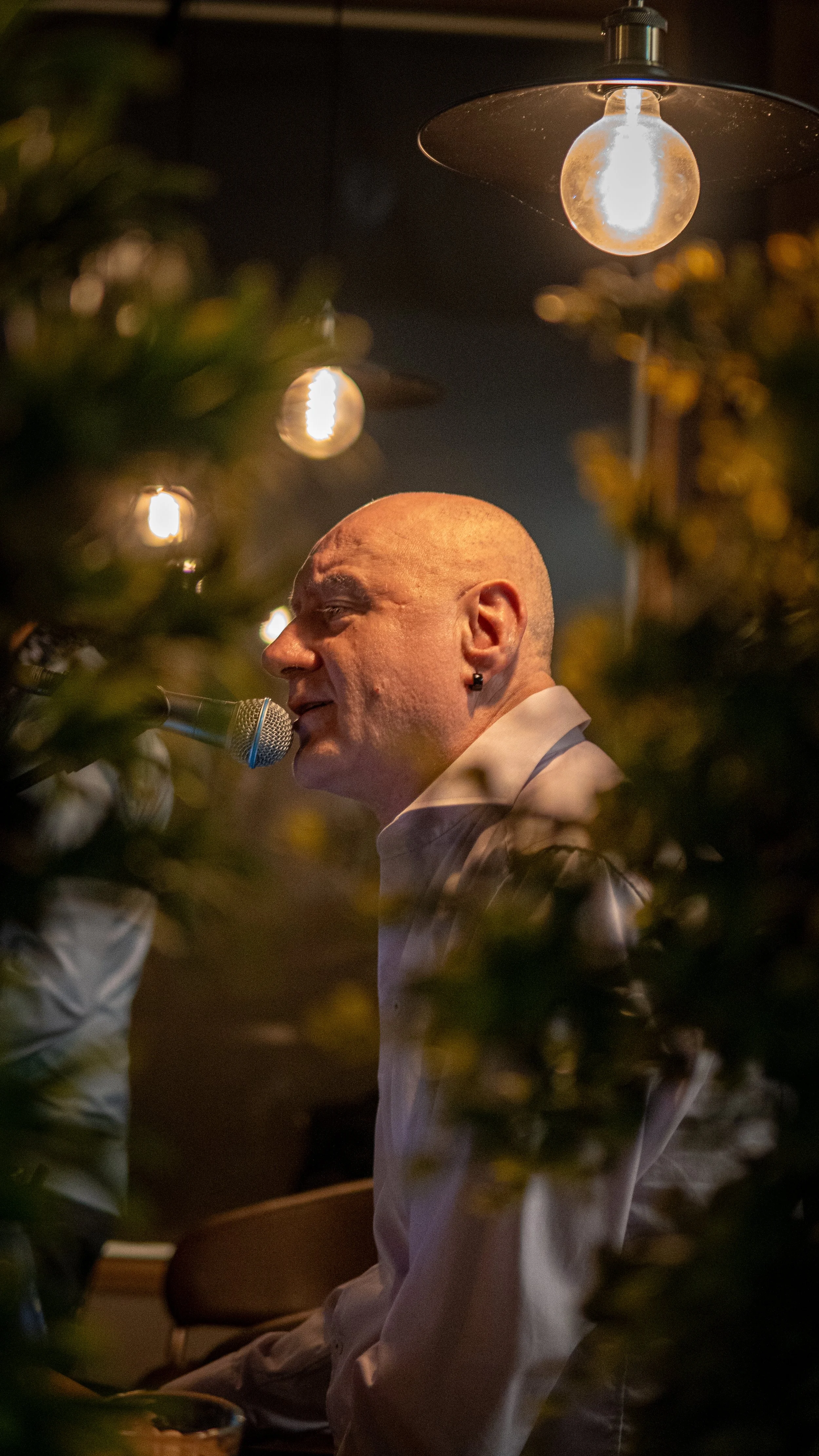 En man med lösögonfransar, cigaretthållare och navelpiercing som spelar piano och sjunger i ett kafé eller restaurang, omgivet av växter och varm belysning.