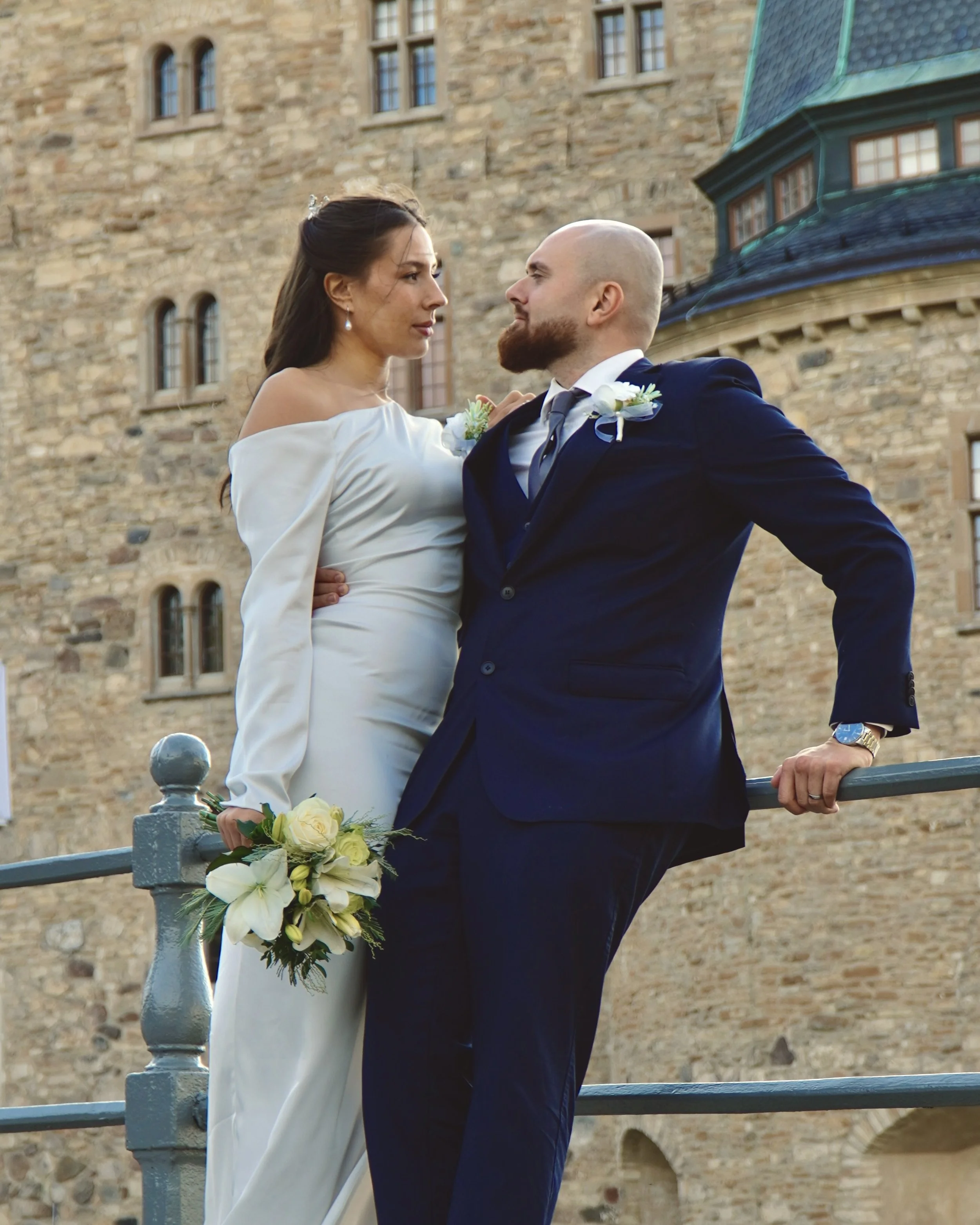 En brud och brudgumme står nära varandra framför ett gammalt stenhus, brudens hand håller en blomsterbukett med vita blommor.