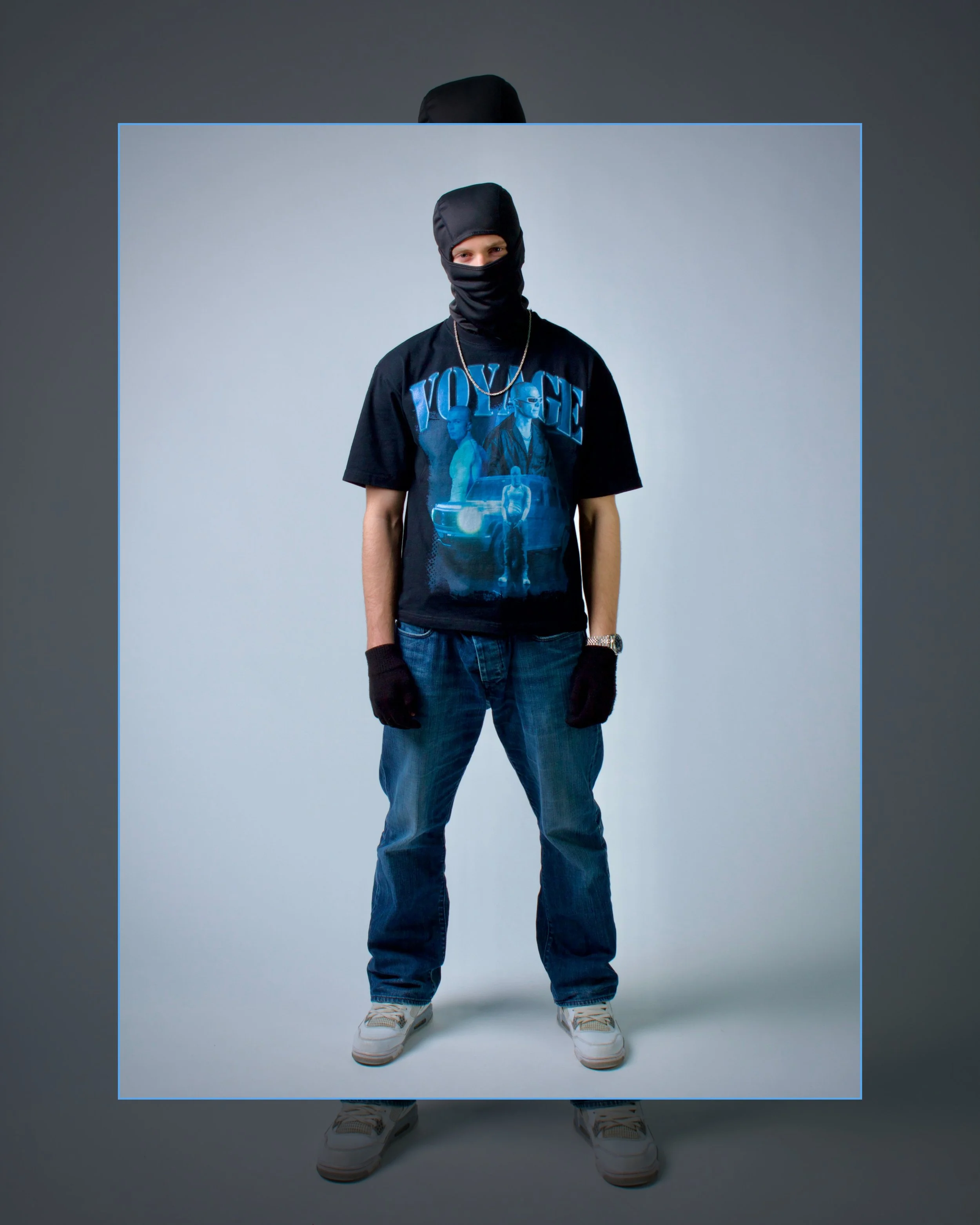 En person klädd i streetwear, inklusive mask, t-shirt med grafik och jeans, står mot en neutral bakgrund.
