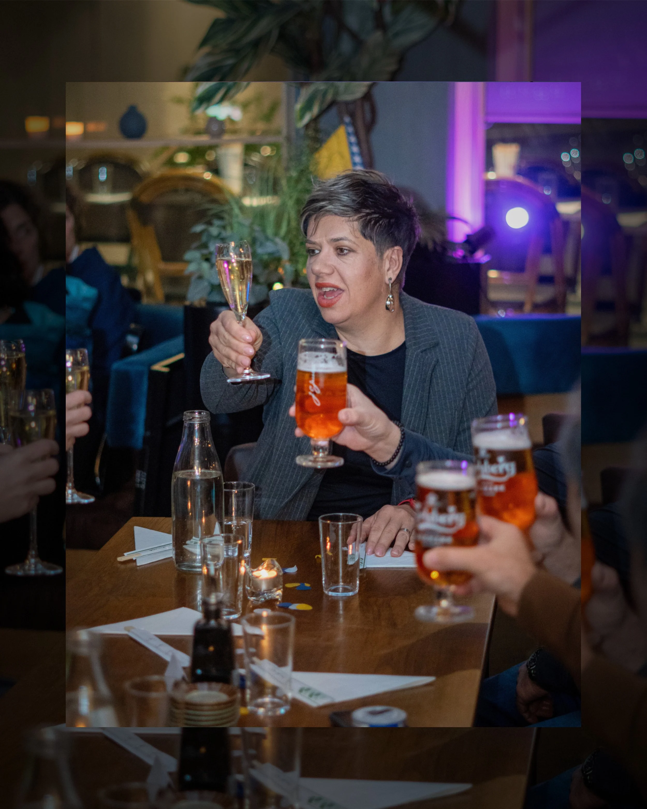 En kvinna med kort mörkt hår och örhängen som håller ett glas champagne och ett glas öl, mitt i ett festligt sällskap vid ett bord.