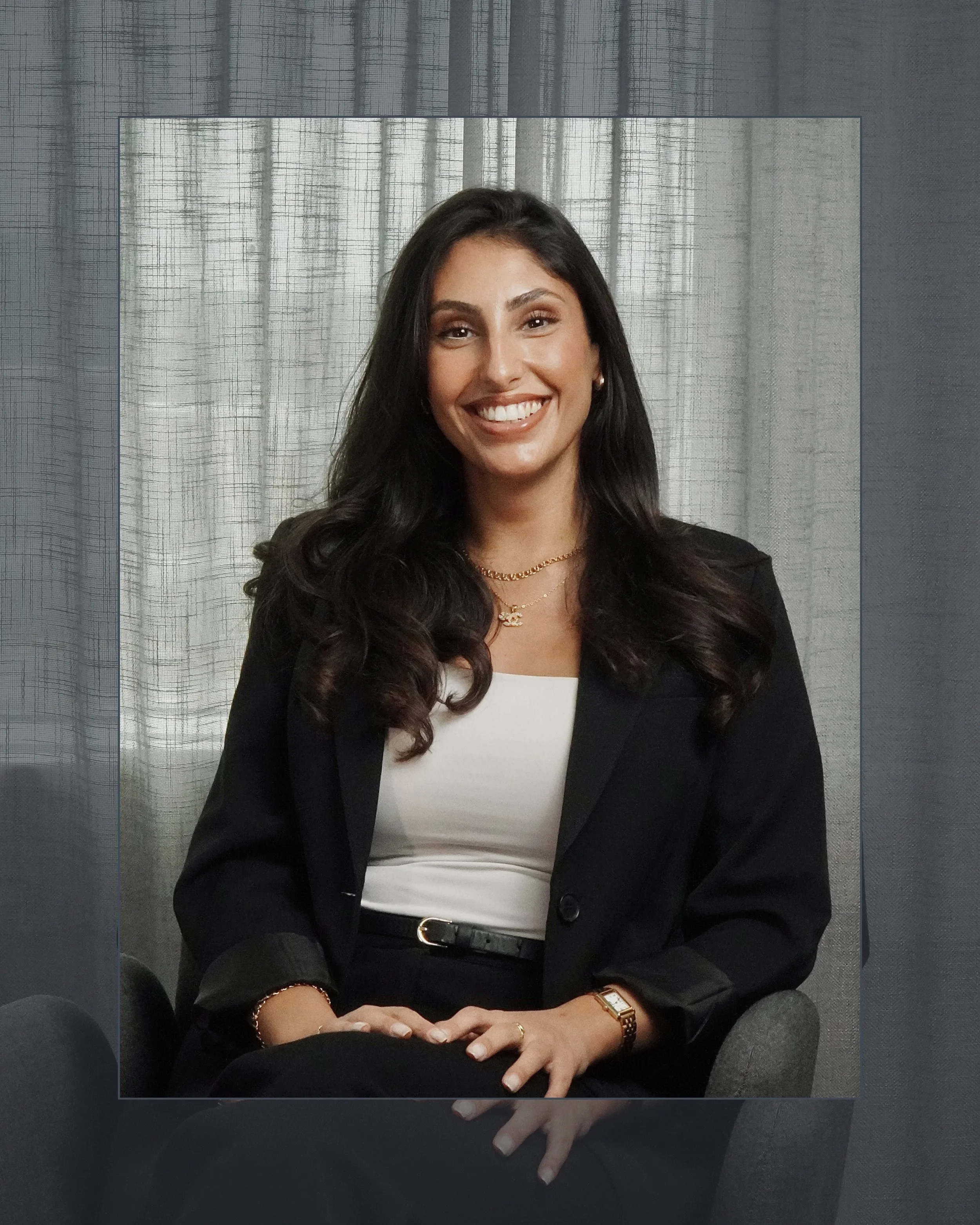 En kvinna med mörkt lockigt hår som ler, sitter i en svart sittyta med en bakgrund av ljusa gardiner.