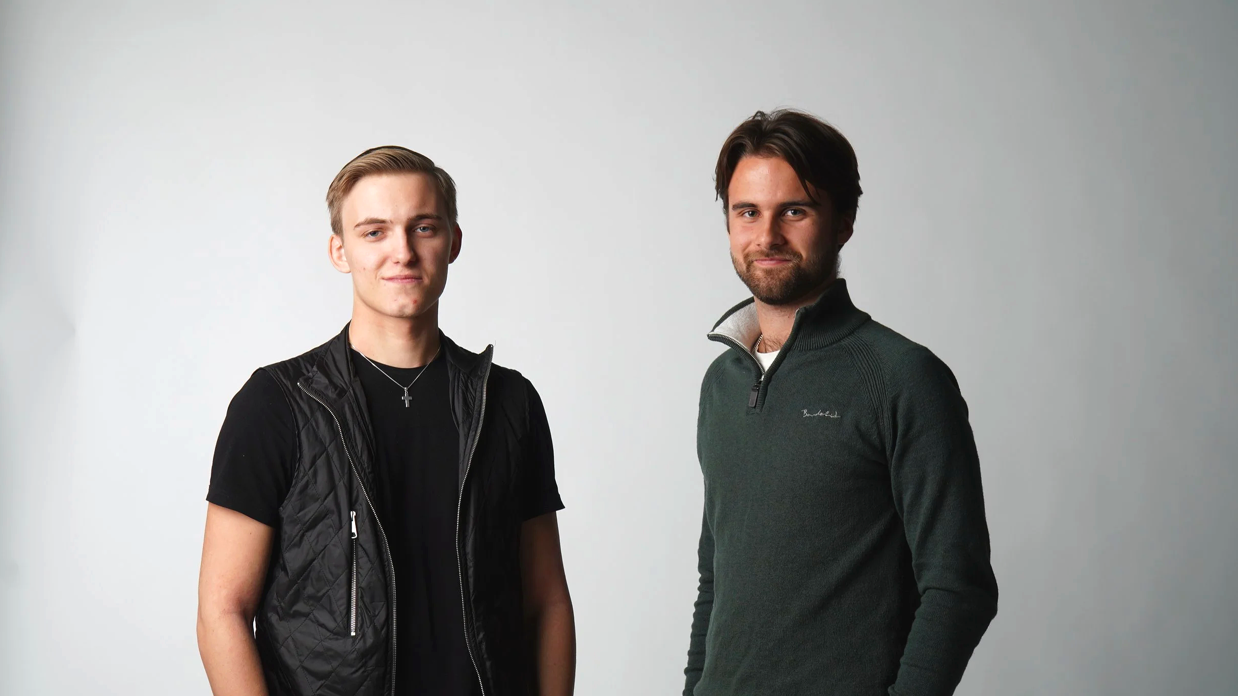 Två män står brett isär och poserar på en ljus bakgrund, båda ser direkt mot kameran och har ett neutralt leende.