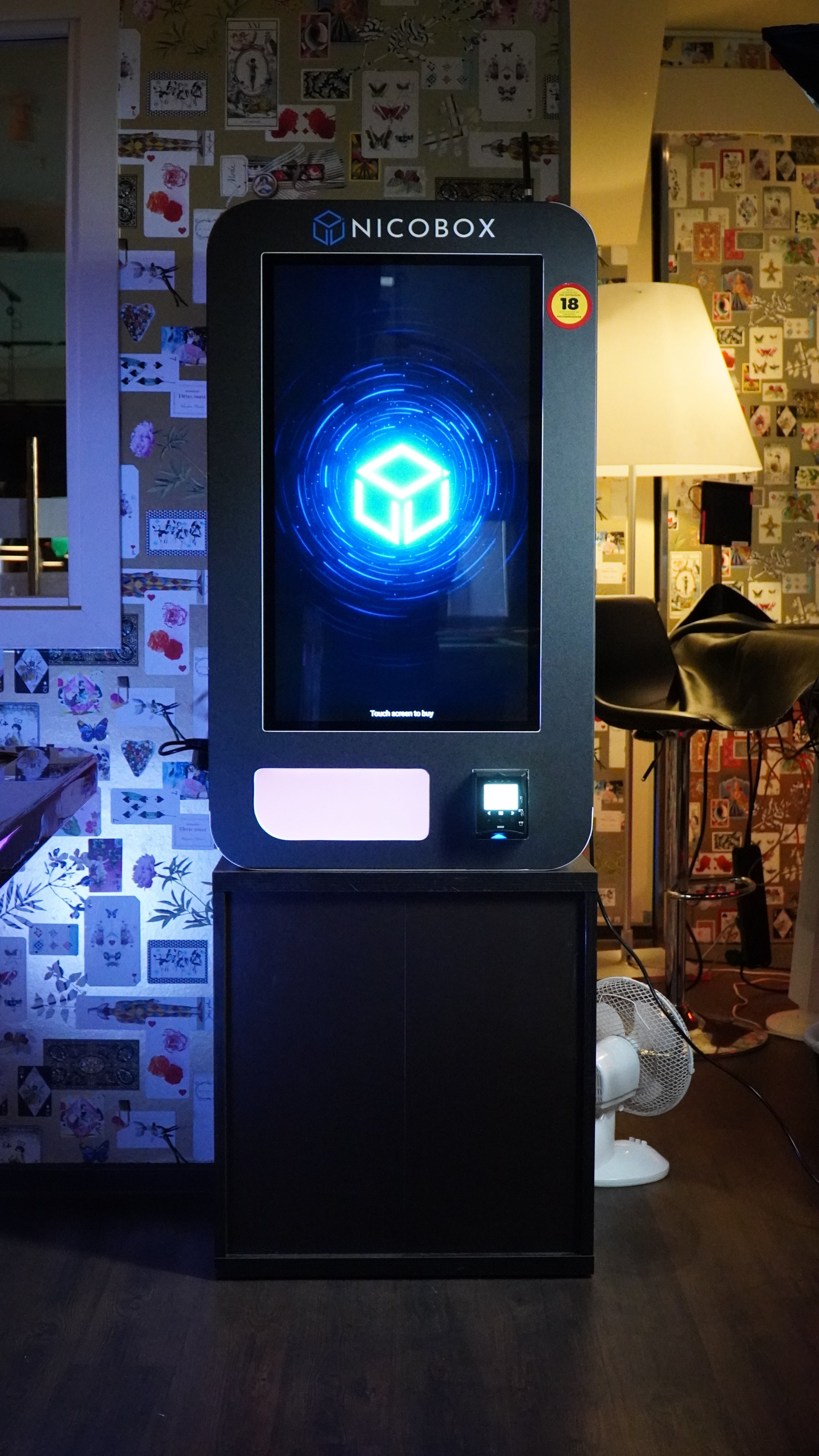 En svart självbetjäningsautomat med en digital skärm och NICOBX-logga, placerad i ett inomhusmiljö med väggdekorationer och möbler i bakgrunden.