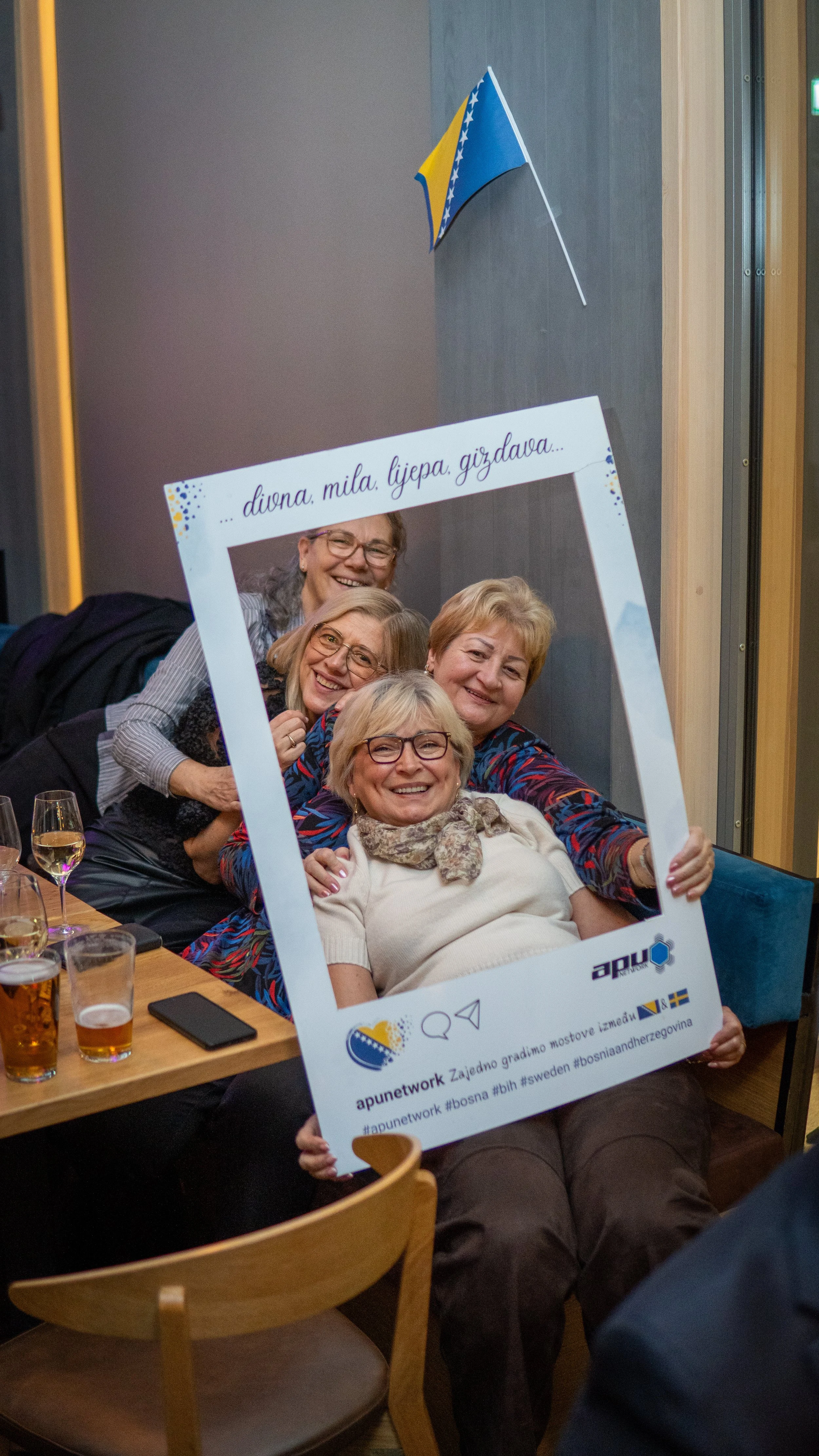 Fem kvinnor som ler och poserar bakom ett fotobås-rekvisita med en svensk flagga och ett textband som säger 'diva, mila, ljepa, gudzava'.