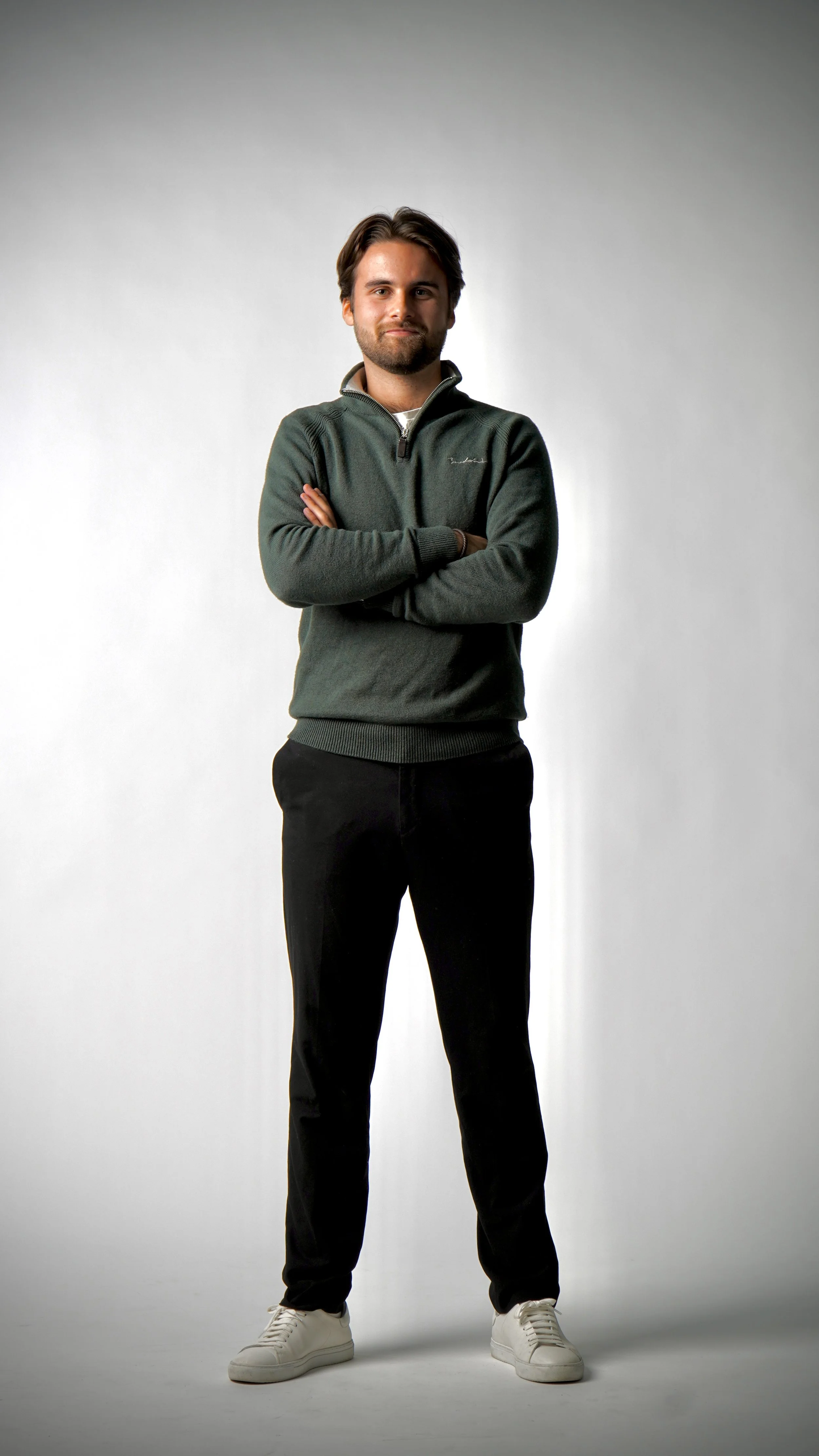 Man står med korsade armar framför en neutral grå bakgrund, klädd i en mörkgrön pullover och svarta byxor.