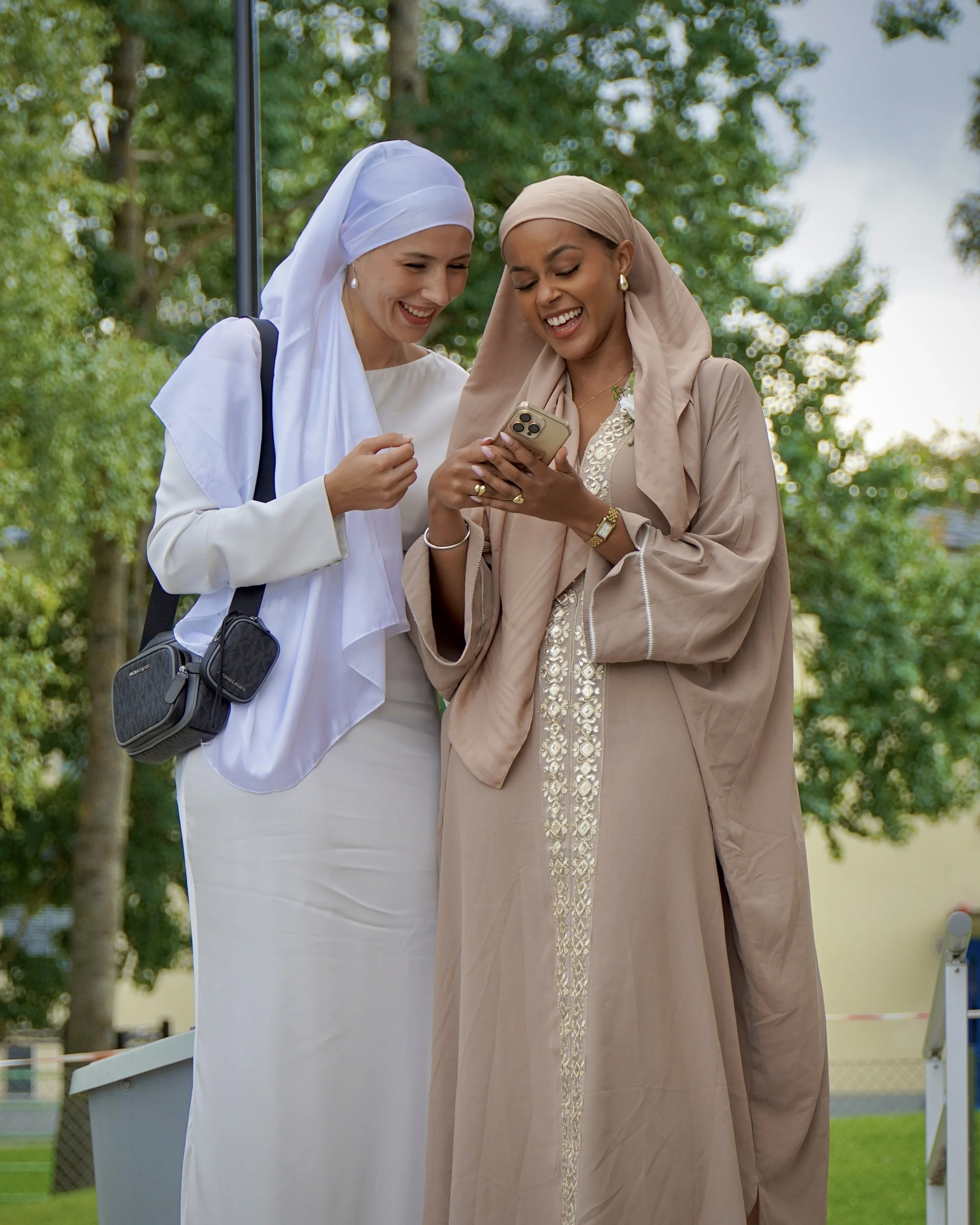 Två kvinnor, båda ler och tittar på en smartphone tillsammans utomhus, med träd i bakgrunden.