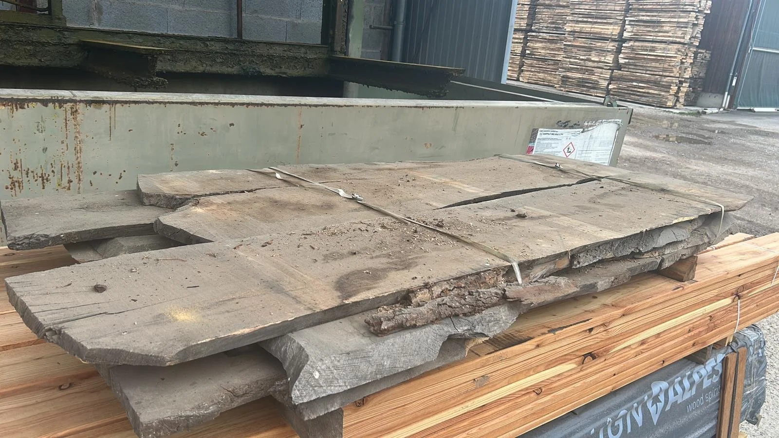 Différentes planches de bois brutes dans un entrepôt ou un espace de stockage. En arrière-plan, un grand conteneur en métal et des piles de bois.