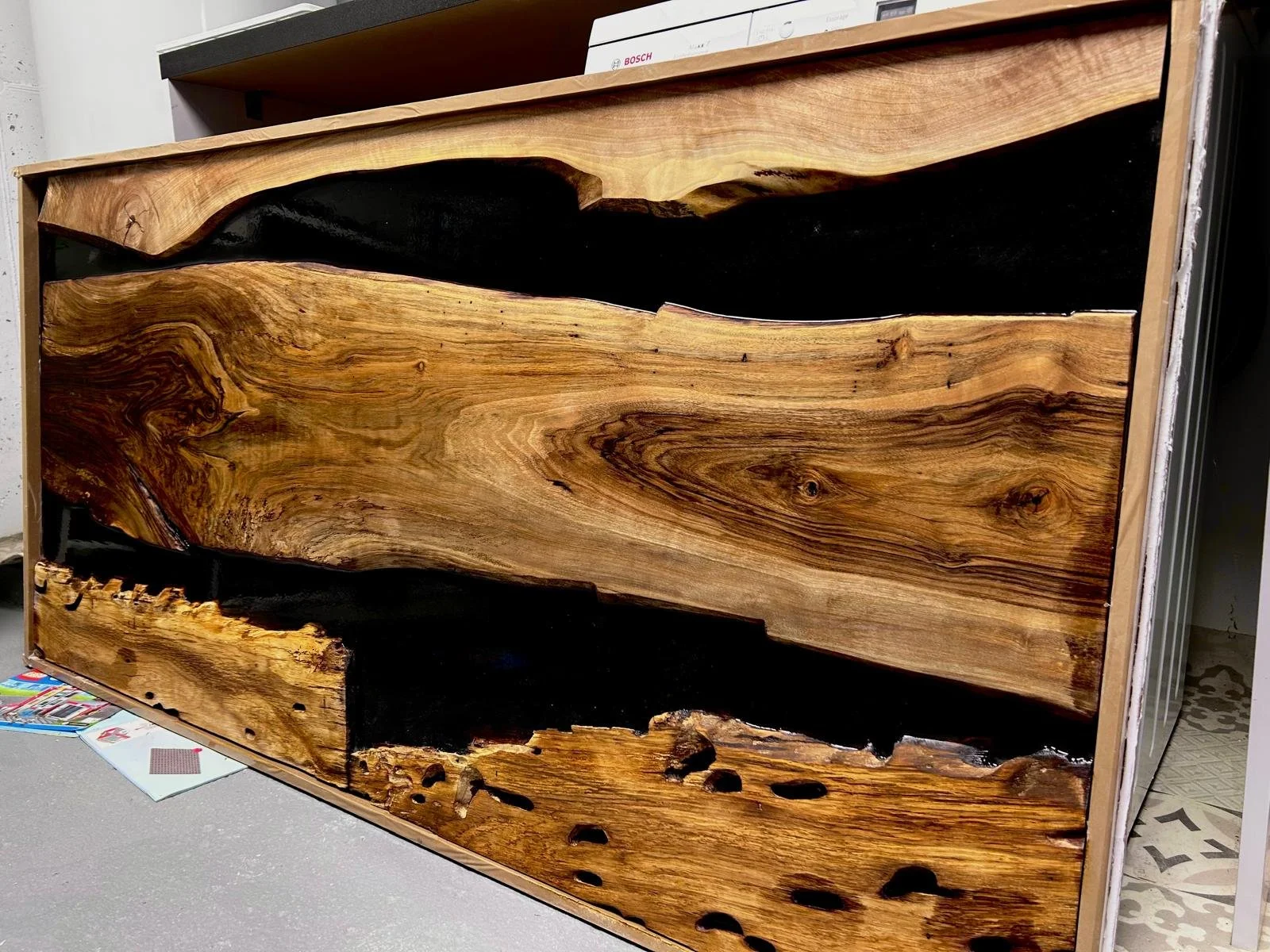 Un meuble en bois avec des sections en bois naturel et une section en résine noire, aspects naturels du bois visible, posé sur une surface en sol gris. 