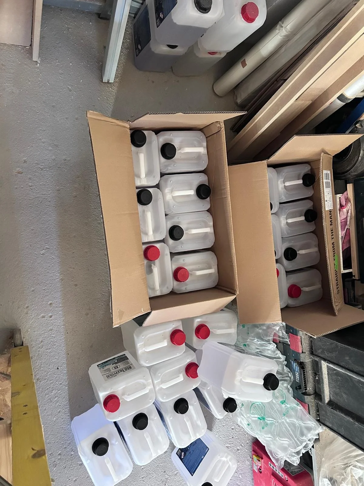 Plusieurs bouteilles en plastique transparent avec des bouchons noirs ou rouges, disposées dans des caisses en carton dans un environnement d'atelier ou de stockage.