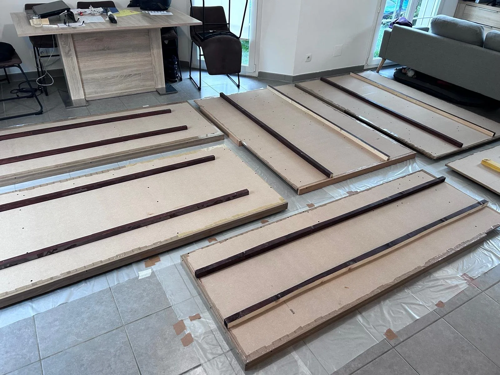 Plusieurs panneaux de bois ou de MDF, avec des bandes de bois foncé, sont disposés sur le sol, en cours de montage ou de fabrication