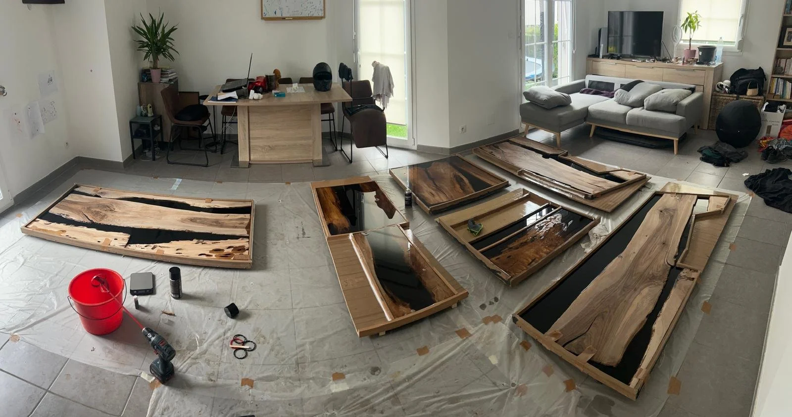 Différentes tables en bois en cours de finition posées au sol dans un salon, avec outils et matériel de travail.