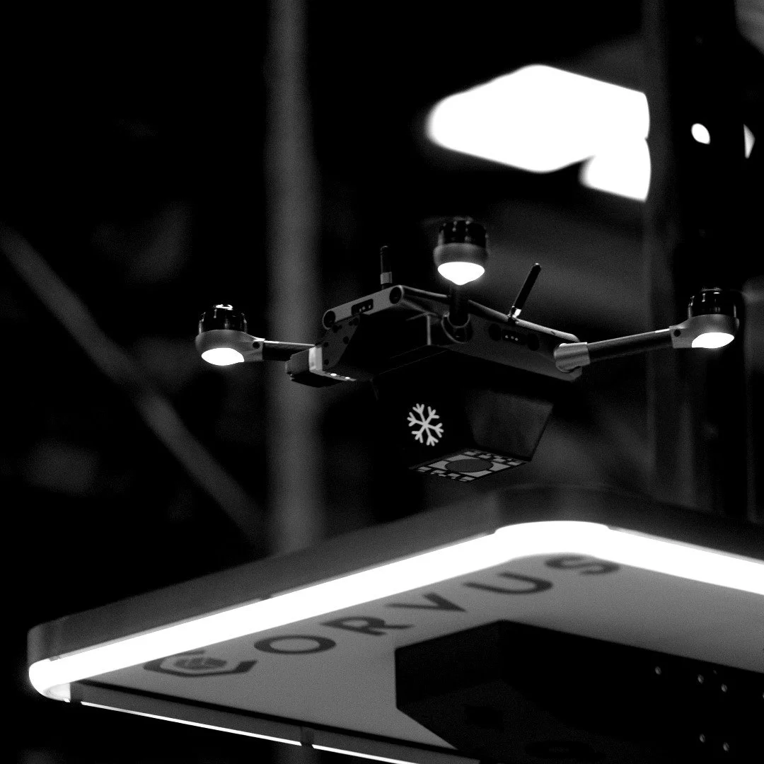 Close-up of a drone hanging from a ceiling, with lights on its arms and a snowflake symbol on its body, against a dark background.