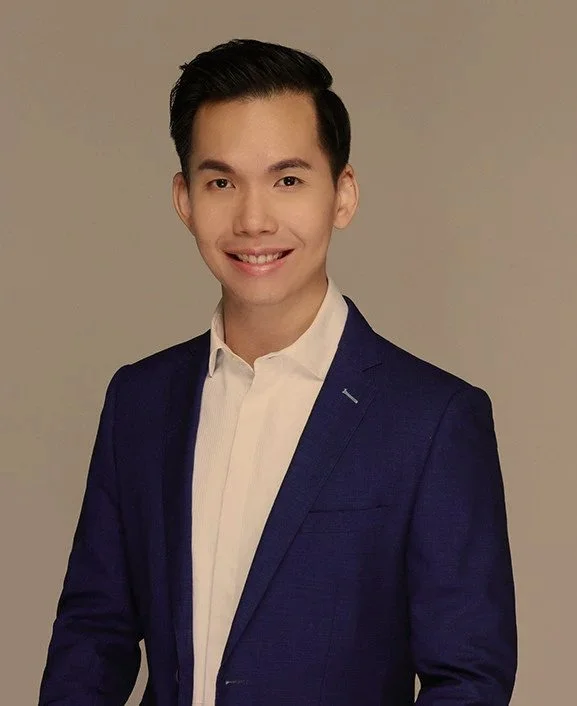 A young man with dark hair smiling, dressed in a white shirt and a dark blue blazer, standing against a plain background.