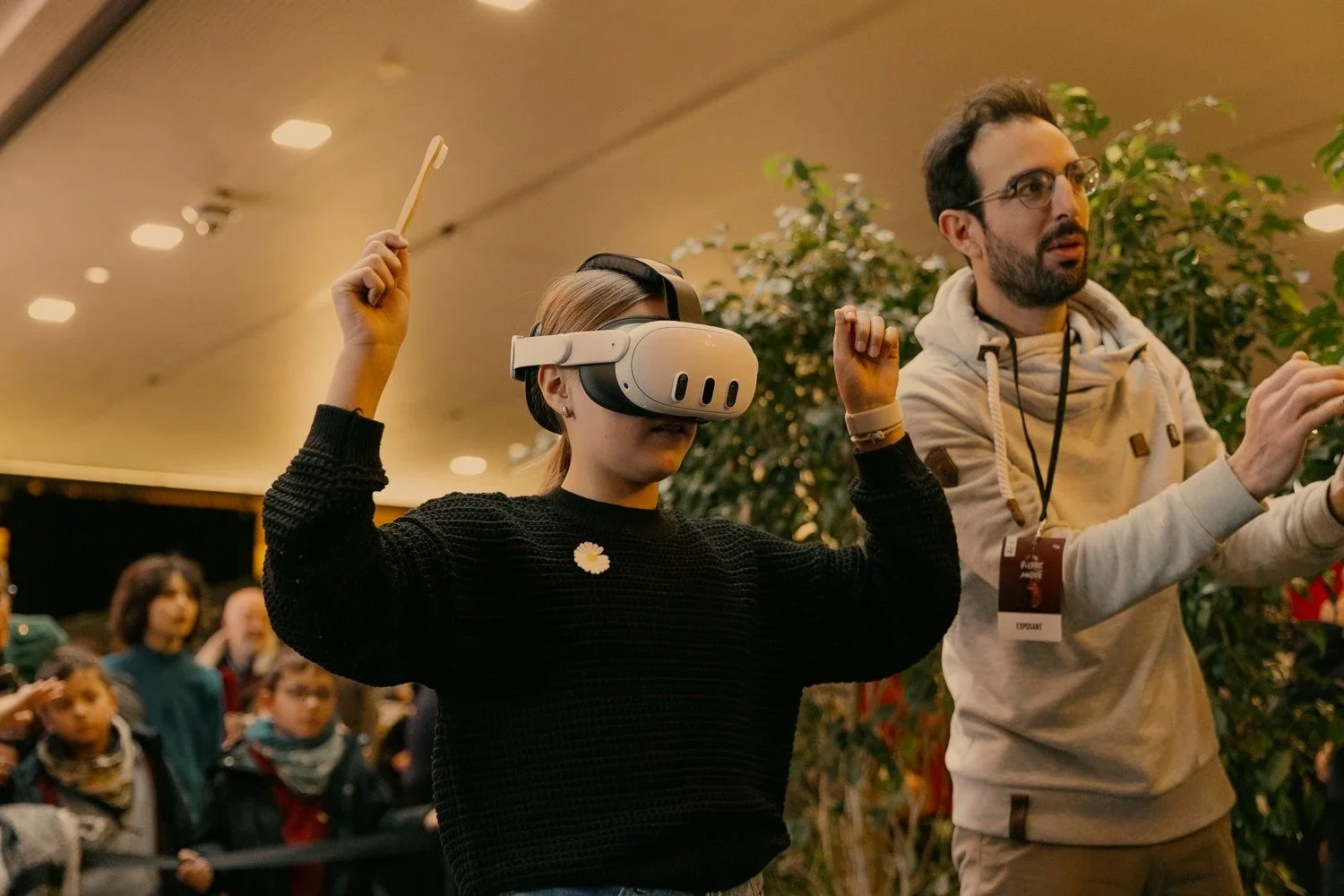 A woman using a virtual reality headset with a crowd of children and a man standing nearby in an indoor setting.