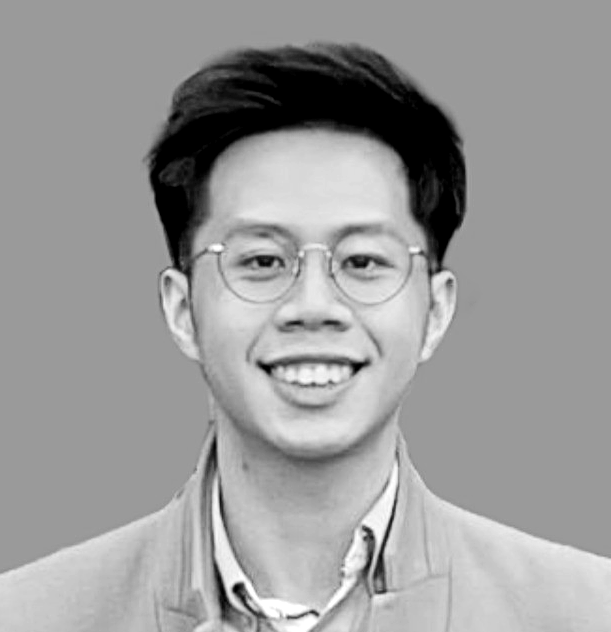 A young man with glasses, smiling, dressed in a suit and tie, against a plain background.