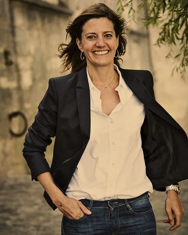 A woman smiling outdoors, wearing a white shirt, black blazer, and jeans.