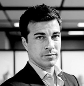 A black and white portrait of a young man with dark hair, wearing a suit and tie, with an office background.