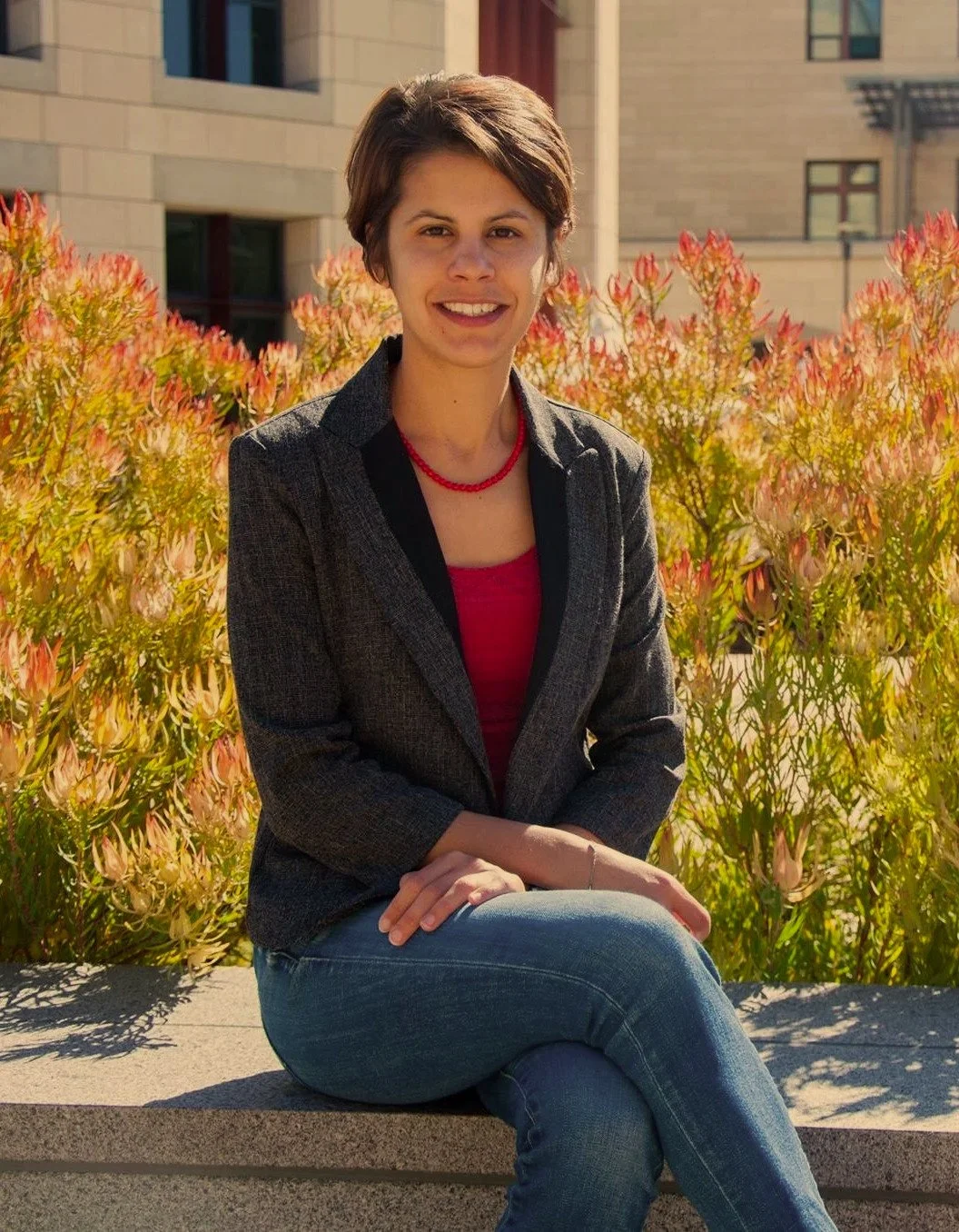 A woman with short brown hair is sitting on a stone bench outdoors, smiling at the camera, with pink and orange flowers and a beige building in the background.