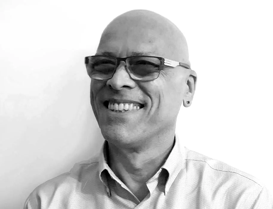 Black and white photo of a smiling, bald man wearing glasses, earrings, and a collared shirt.