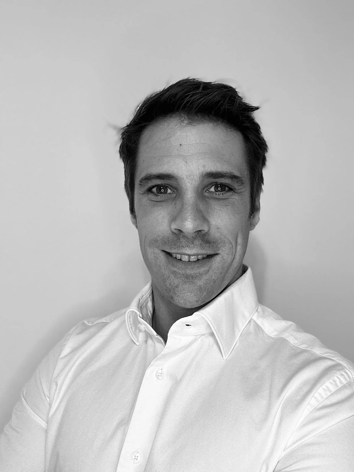 Black and white photo of a man smiling, wearing a collared shirt, standing against a plain background.