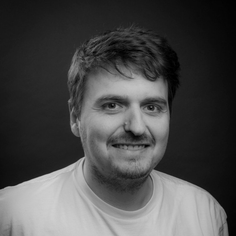 Black and white portrait of a smiling man with short, slightly messy hair, wearing a light-colored t-shirt, against a solid dark background.