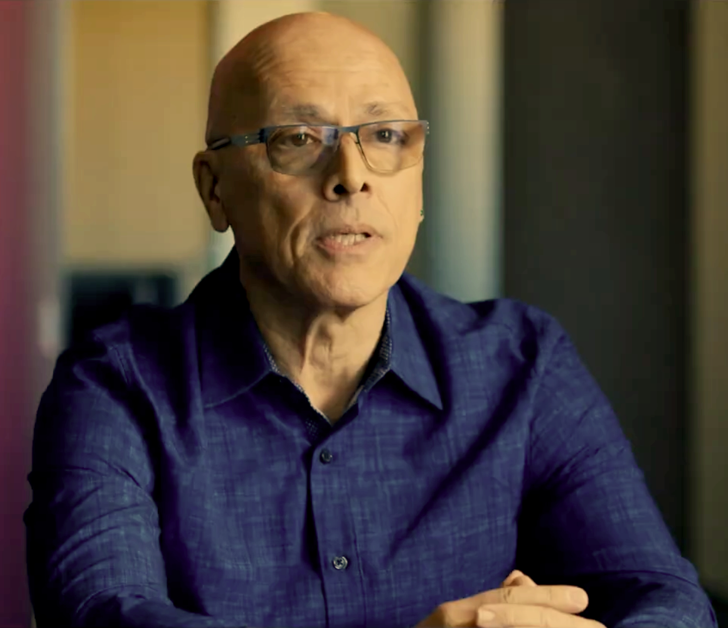 An older man with glasses and a bald head, wearing a dark blue shirt, speaking during an interview or meeting in an indoor setting.