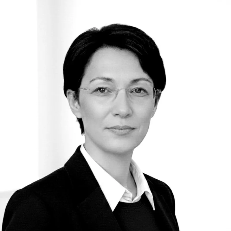 Black and white professional portrait of a woman with short hair, glasses, and a serious expression, wearing a suit jacket and white shirt.