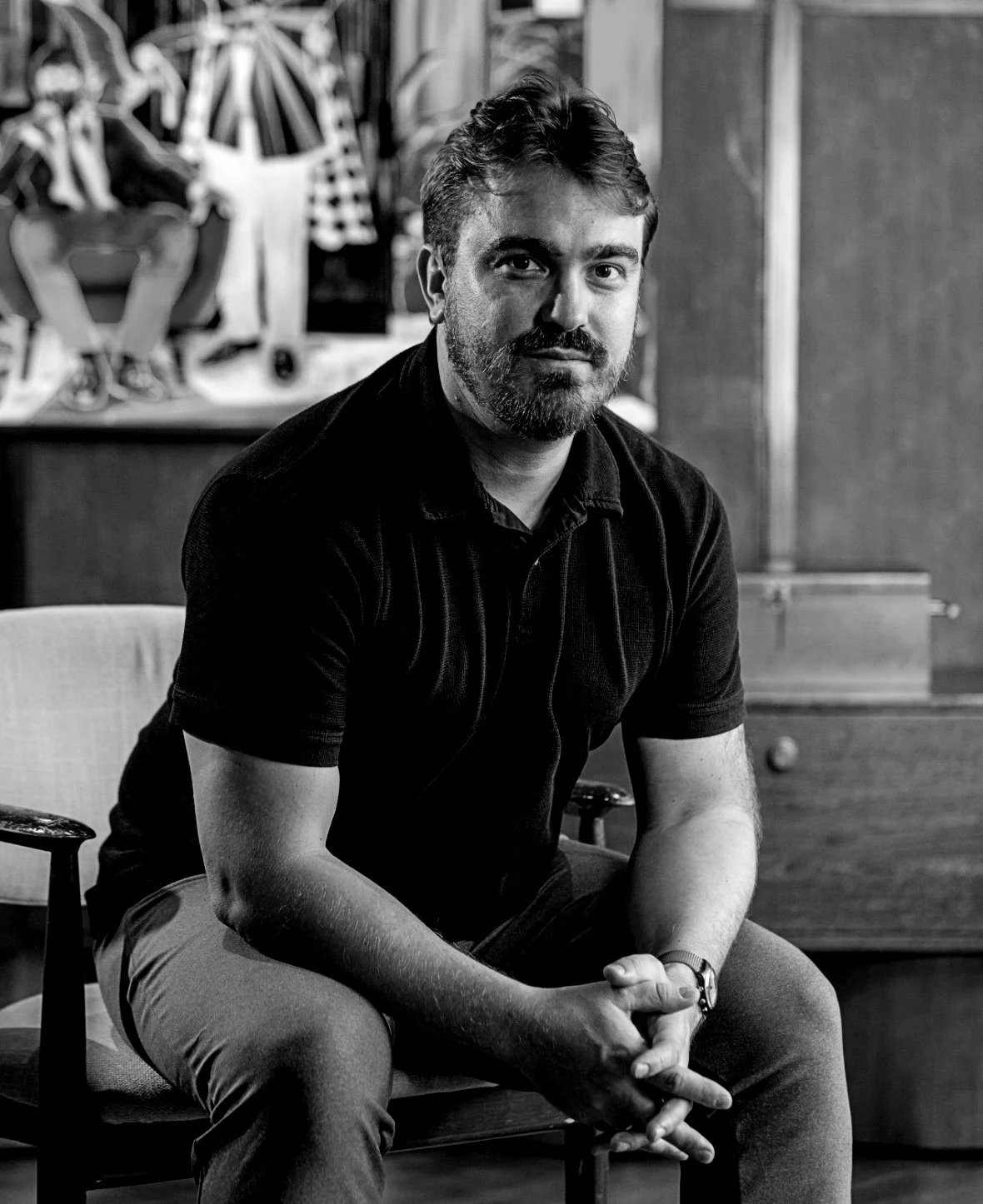 A man with a beard and short hair sitting on a chair in a room with artwork in the background.