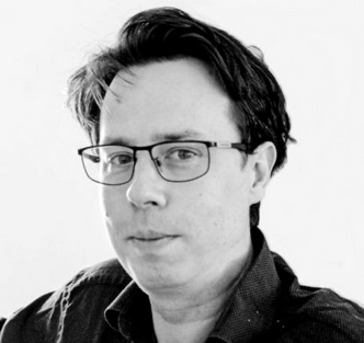 Black-and-white portrait of a person with dark, wavy hair, glasses, and a serious expression, wearing a dark collared shirt.