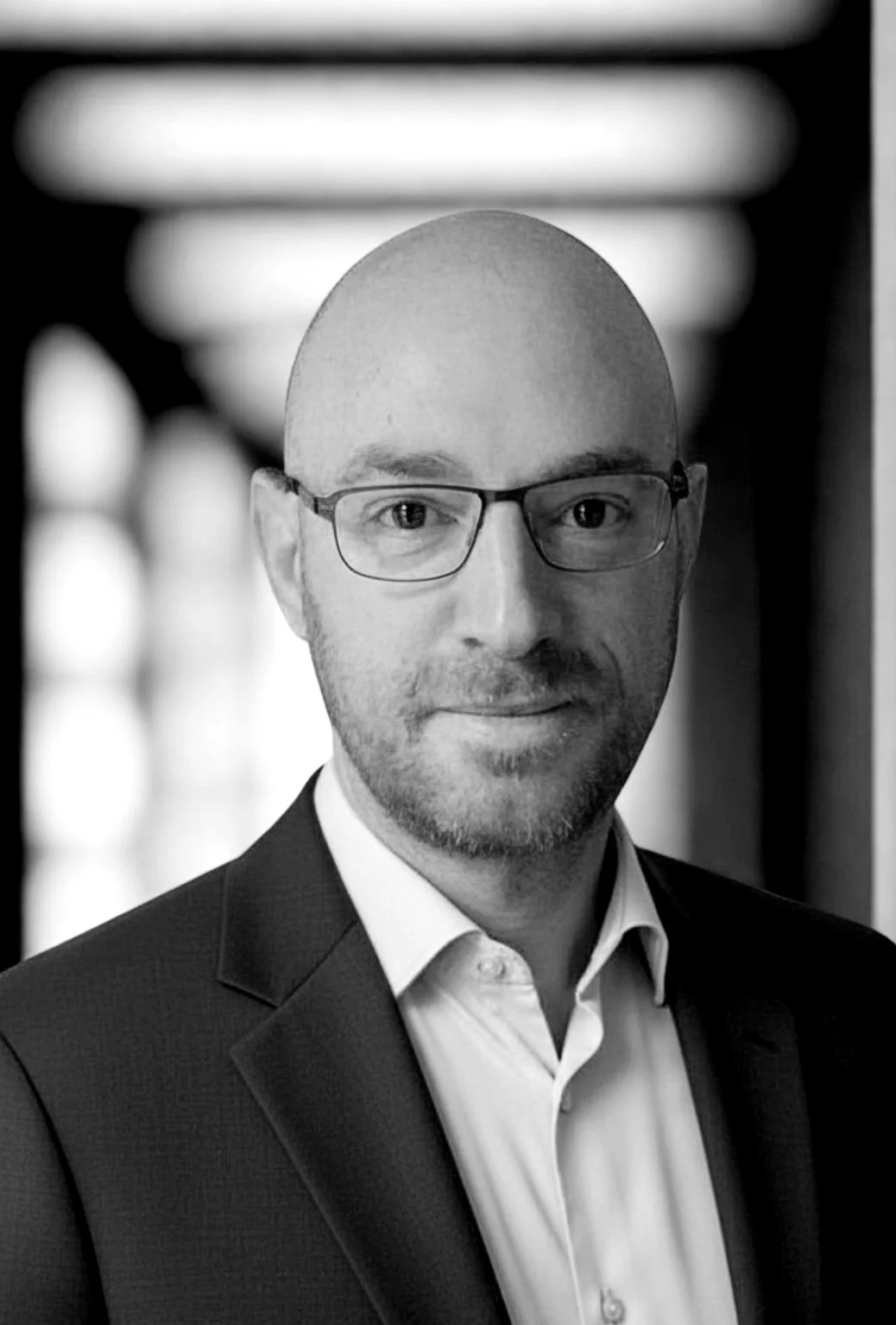 Black and white professional portrait of a bald man with glasses, wearing a suit and white shirt, in an indoor setting with blurred background.