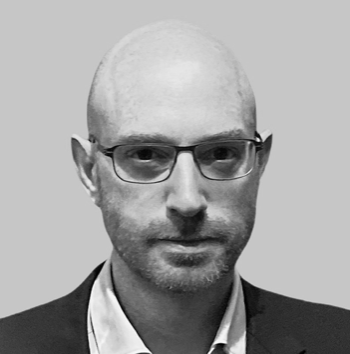 A black and white portrait of a man wearing glasses, a collared shirt, and a blazer.