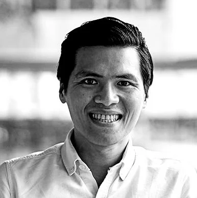 A smiling man with short, dark hair in a white button-up shirt, standing indoors with large windows in the background.