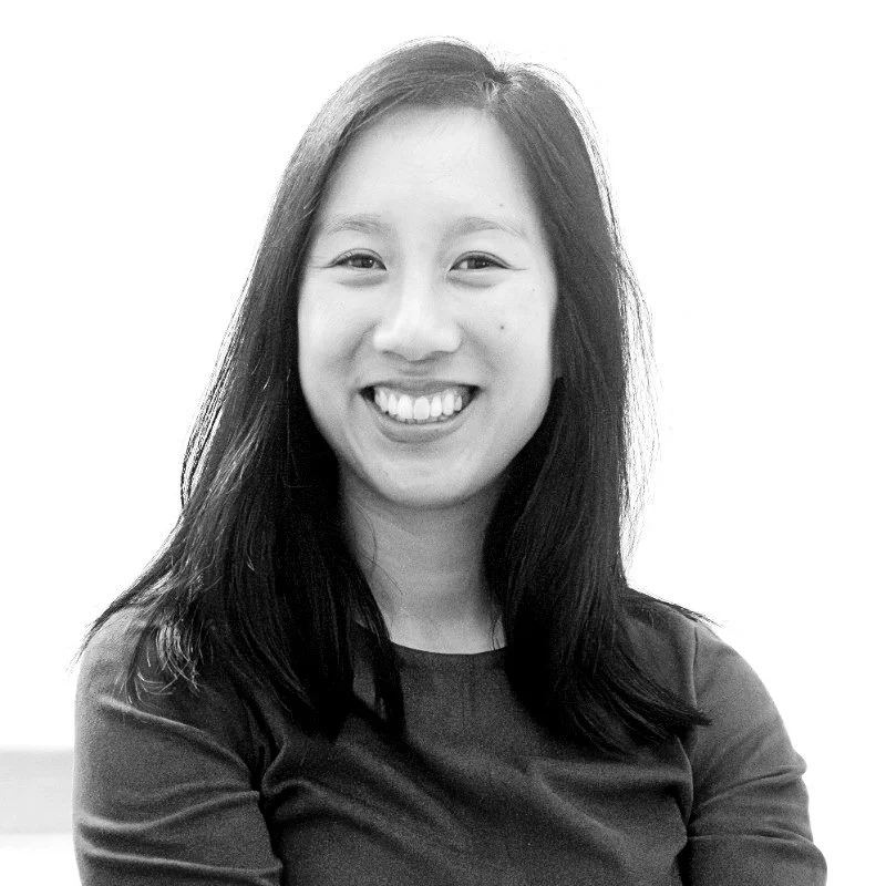Black and white portrait of a smiling woman with long dark hair, wearing a dark top, with a plain light background.