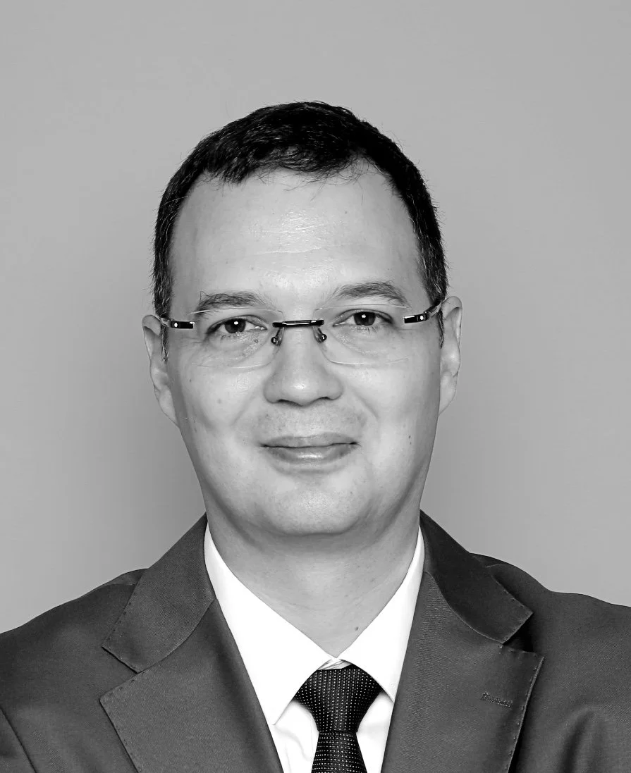 Black and white professional portrait of a man in a suit with glasses, smiling, against a plain background.