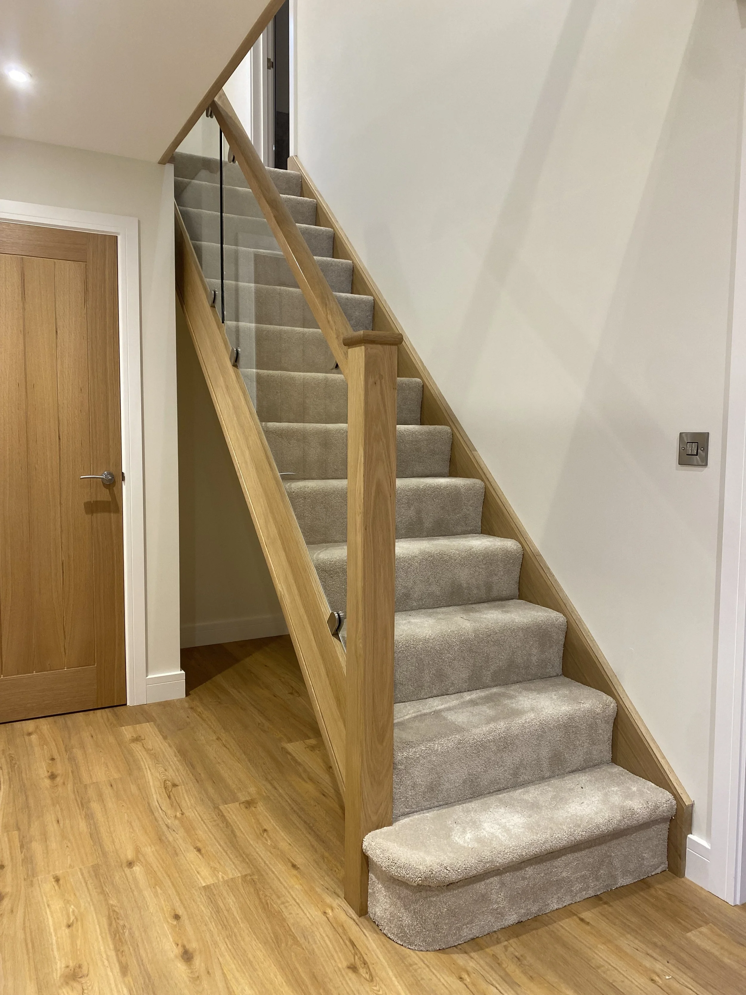 Interior view of a staircase with beige carpet, wooden banister, and light-colored walls, next to a wooden door on the left.