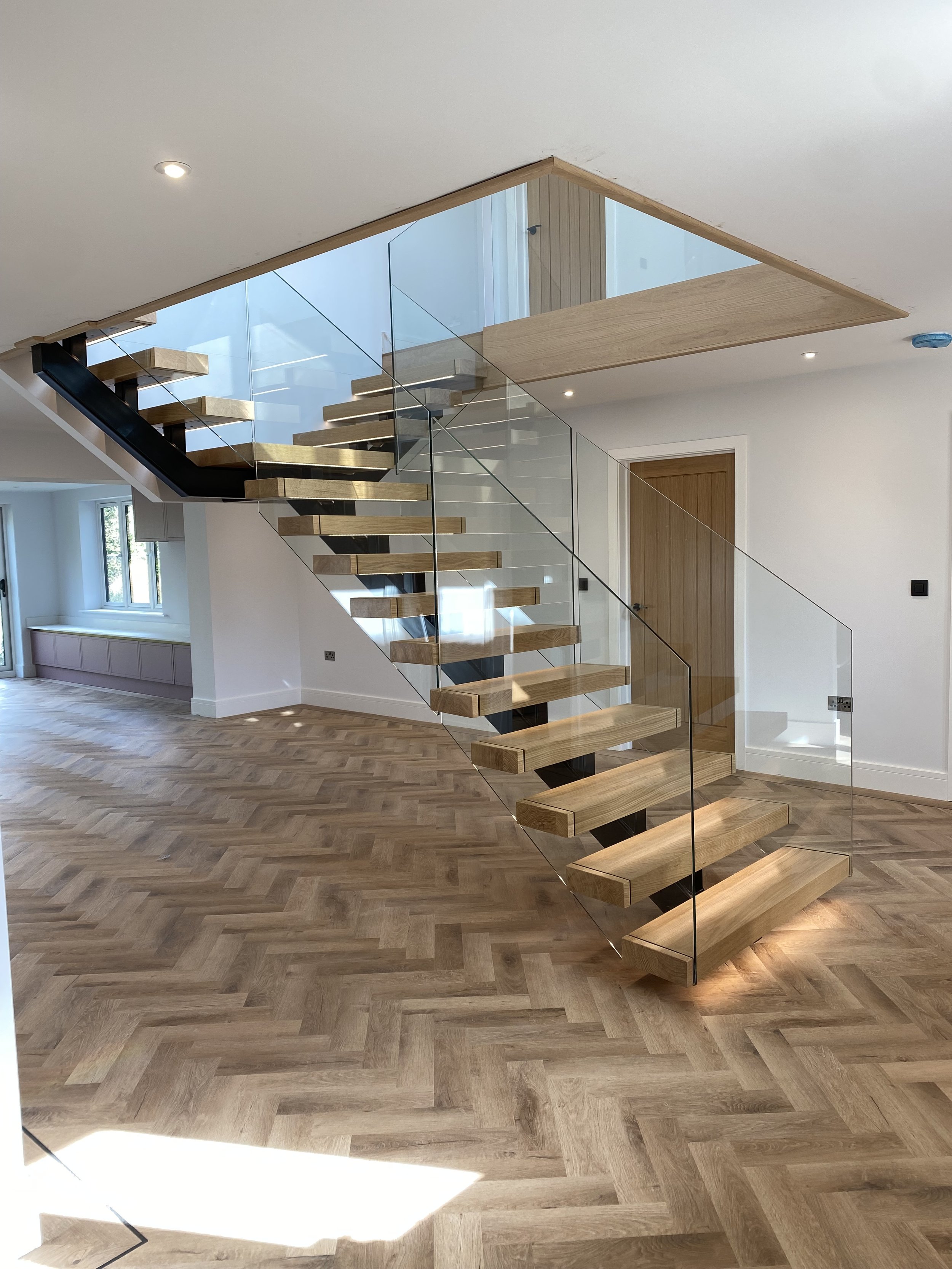 Modern interior staircase with wooden steps, glass railing, and open space with wooden flooring and white walls.