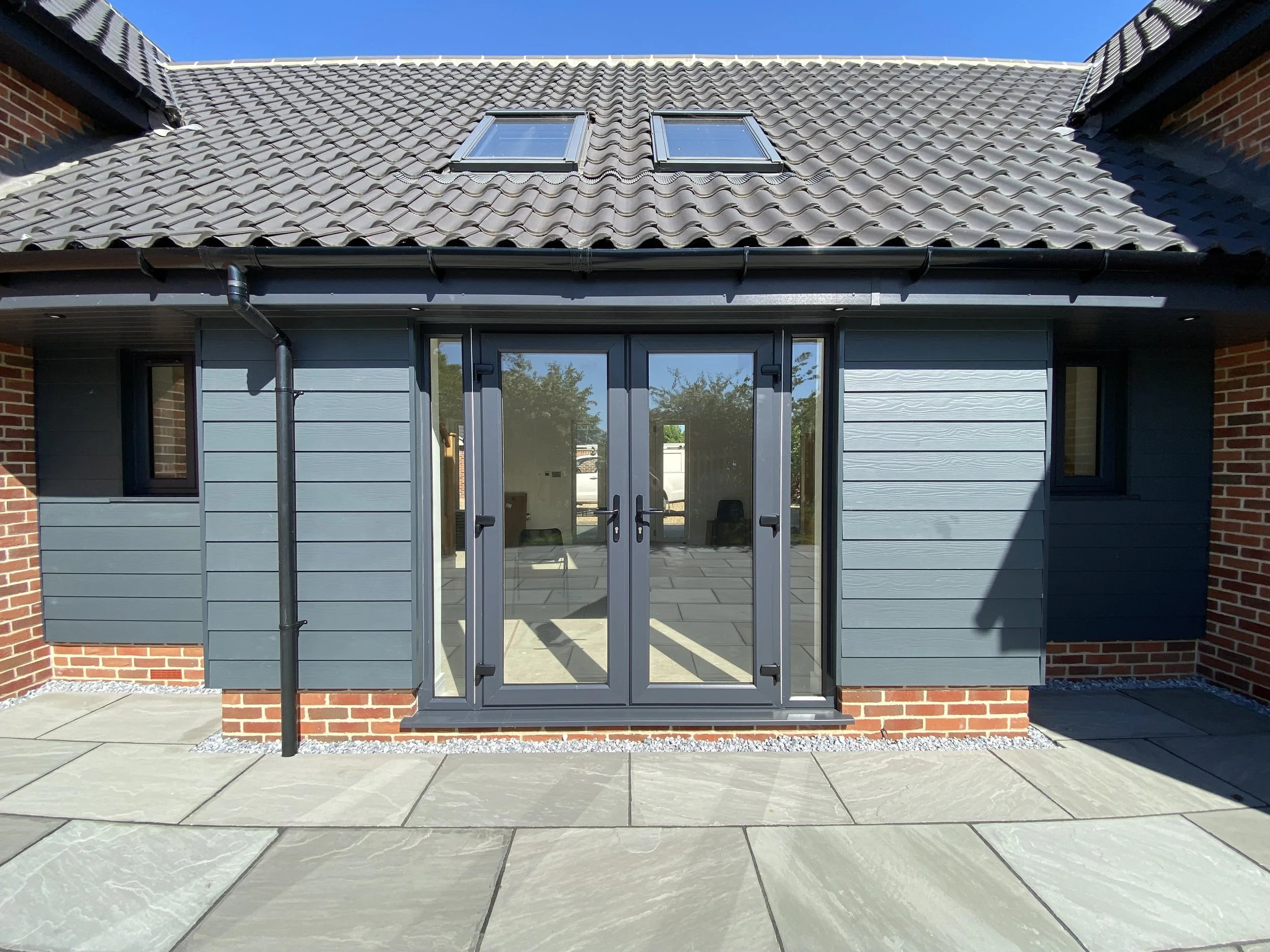 Rear patio of a house with gray sliding glass door, blue siding, small window, brick foundation, and tiled roof with skylights.
