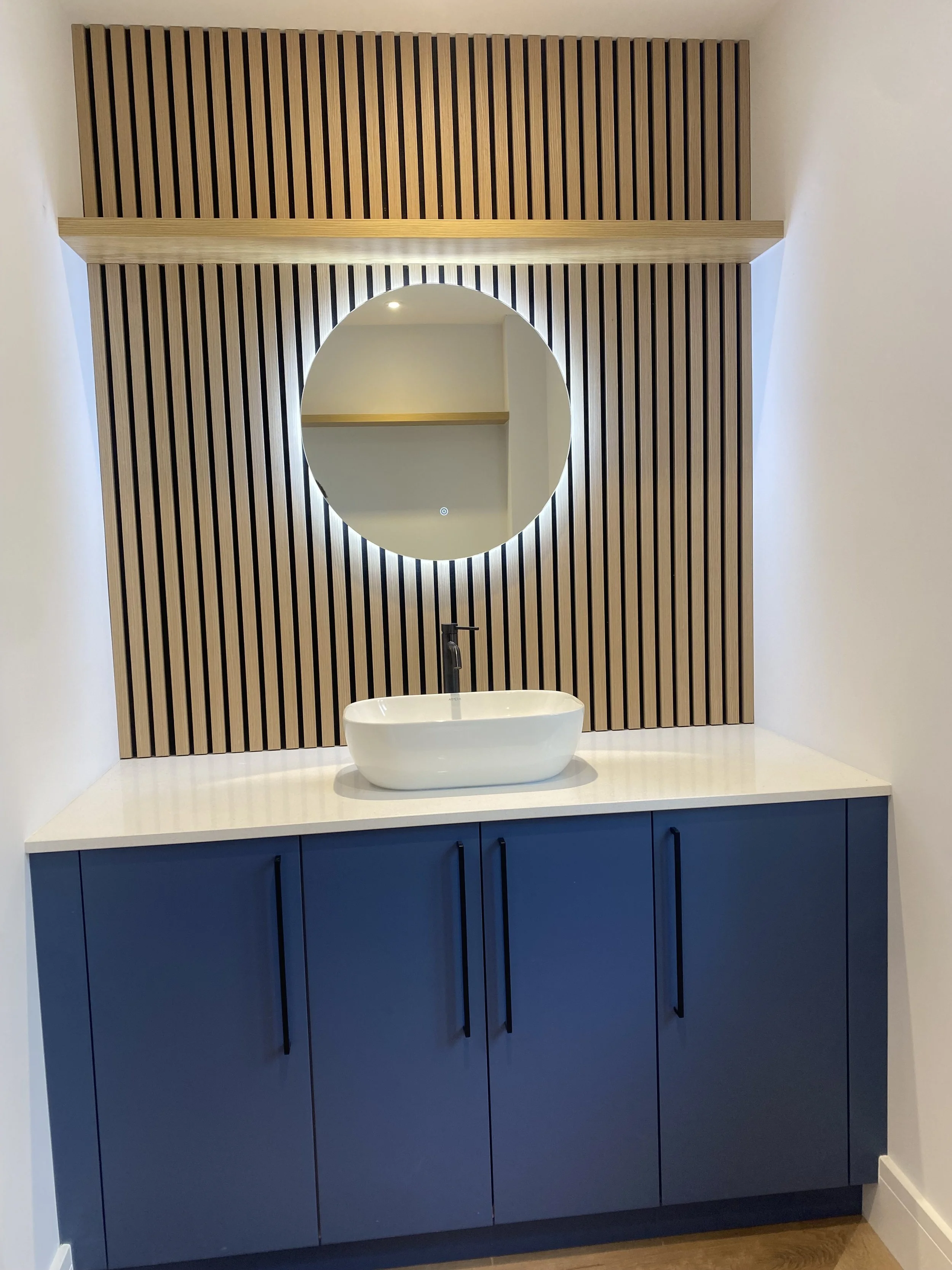 Modern bathroom vanity with blue cabinets, white countertop, oval vessel sink, black faucet, round backlit mirror, and wooden slatted wall paneling.